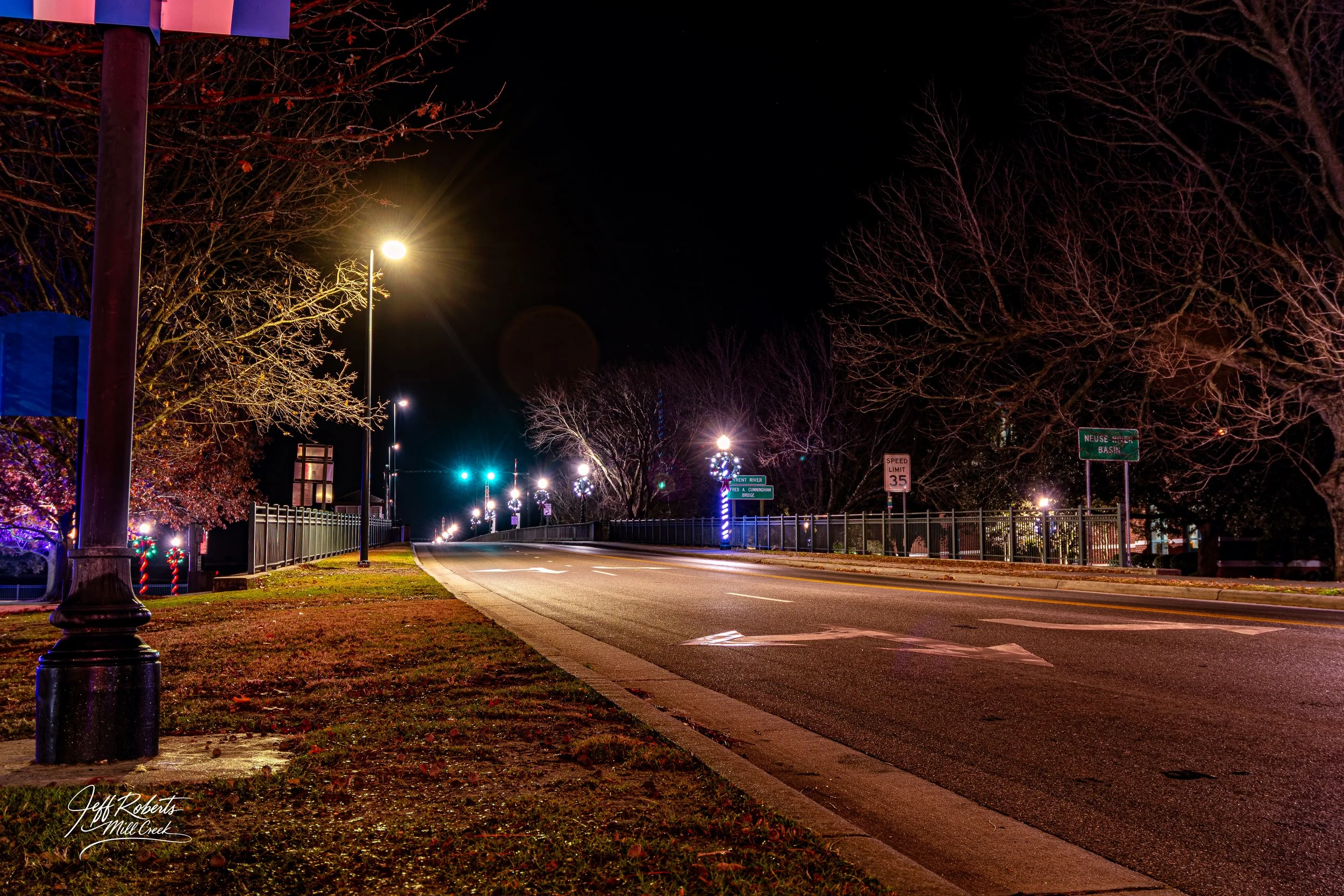 Nighttime street scene with lit street lamps, bare trees, a fenced sidewalk, and holiday decorations on a clear night.