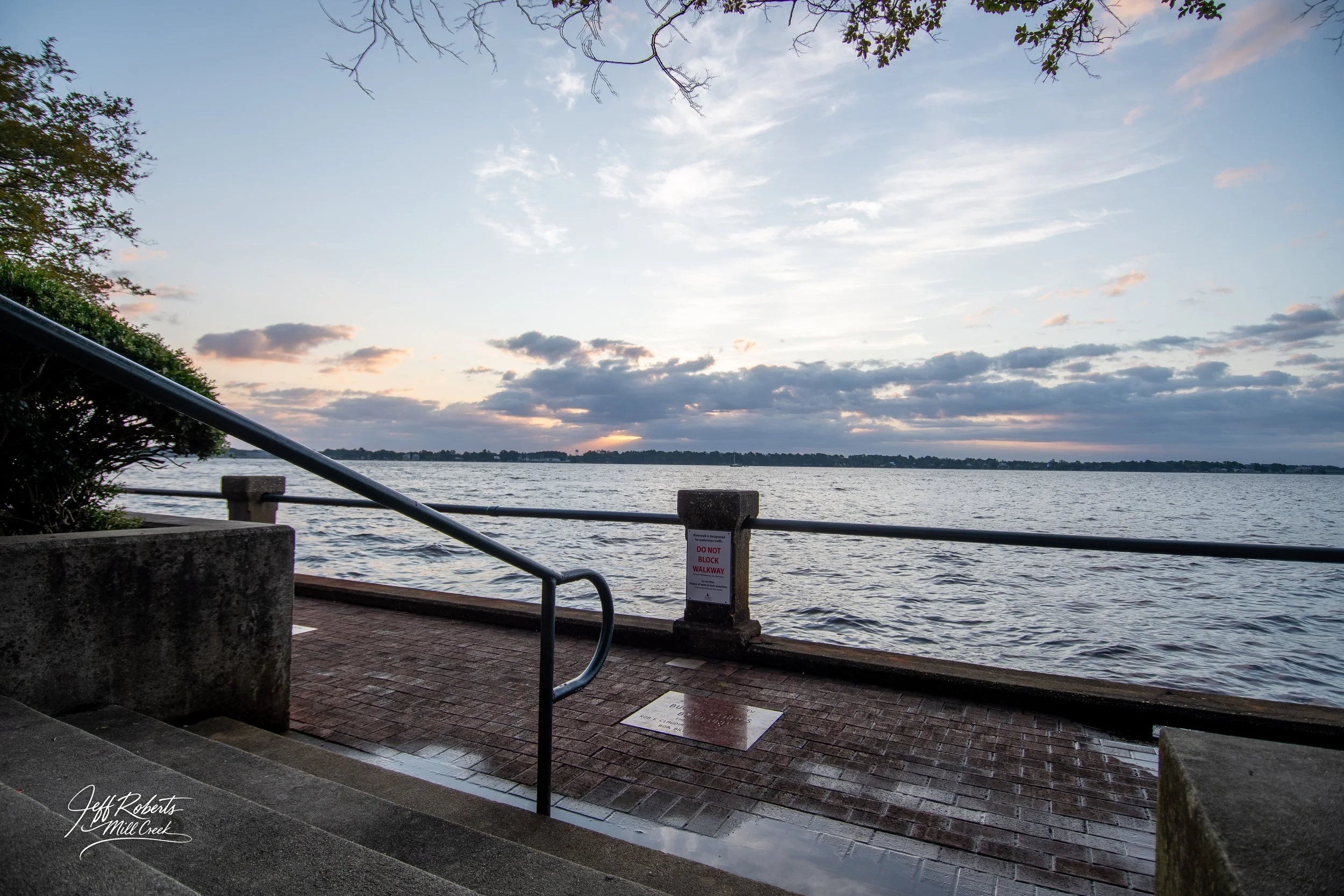 A lakeside walkway with a metal railing, wet brick pavement, and a 'Do Not Block Walkway' sign, overlooking a calm body of water under a partly cloudy sky at sunset.