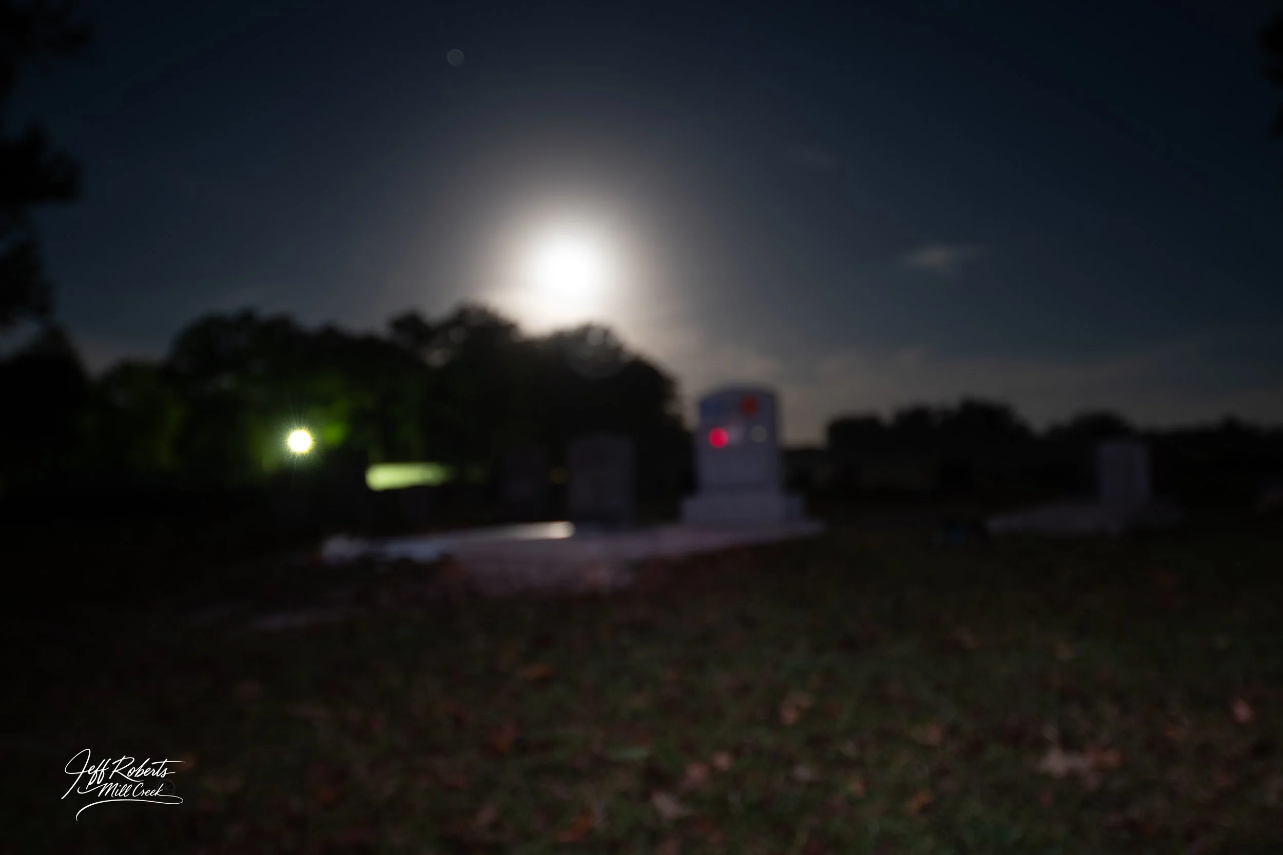 A nighttime scene at a cemetery with tombstones, illuminated by moonlight, with trees in the background and a glow from distant lights.