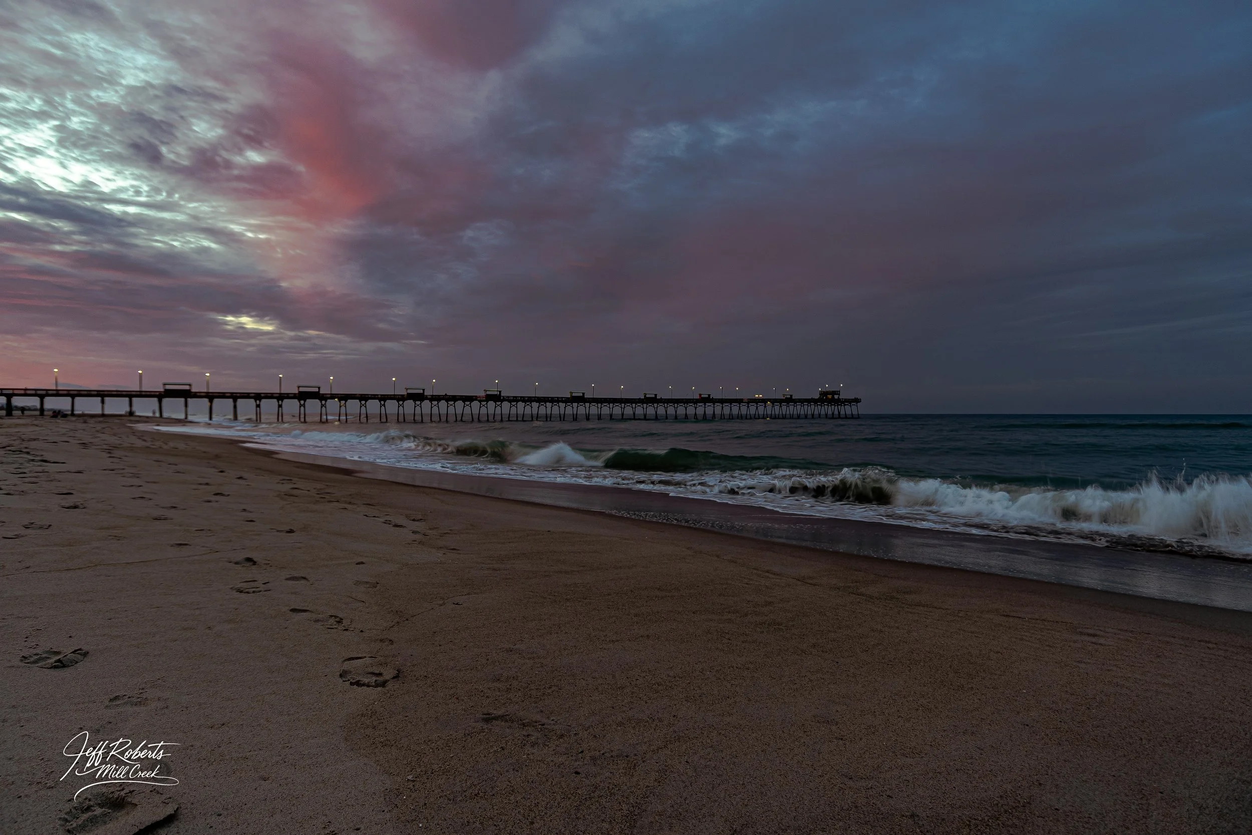 A beach at dusk with footprints in the sand, and a pier extending into the ocean under a partly cloudy sky with pink and purple hues.