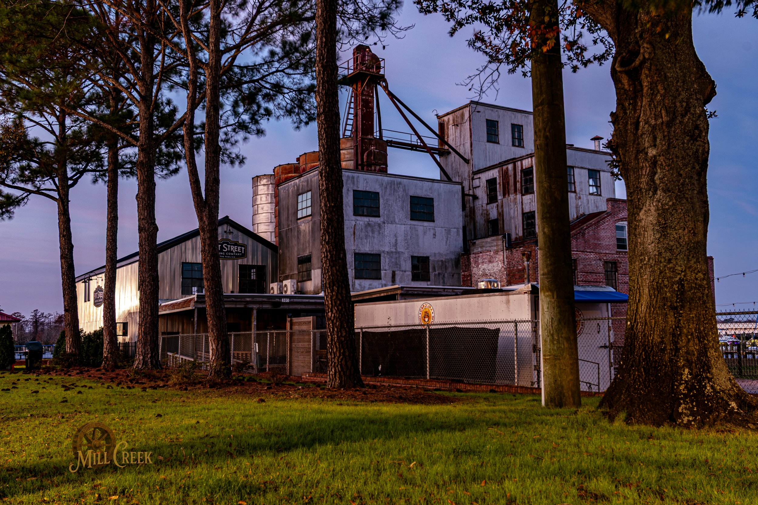 Old industrial building surrounded by trees, with a grassy lawn in the foreground and a fence around the area.