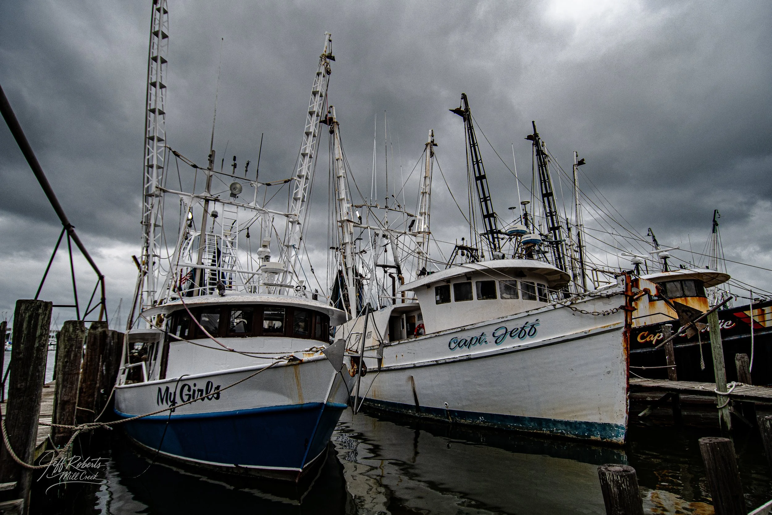 Two boats docked at a harbor under dark, stormy clouds, with one named 'My Girls' and the other 'Capt. Debo'.