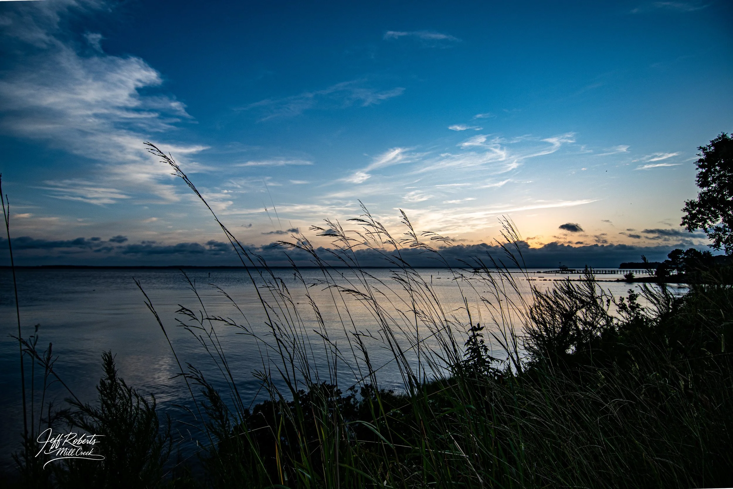 A serene river scene at sunset with tall grasses in the foreground, a calm river, and a sky filled with clouds and fading sunlight.