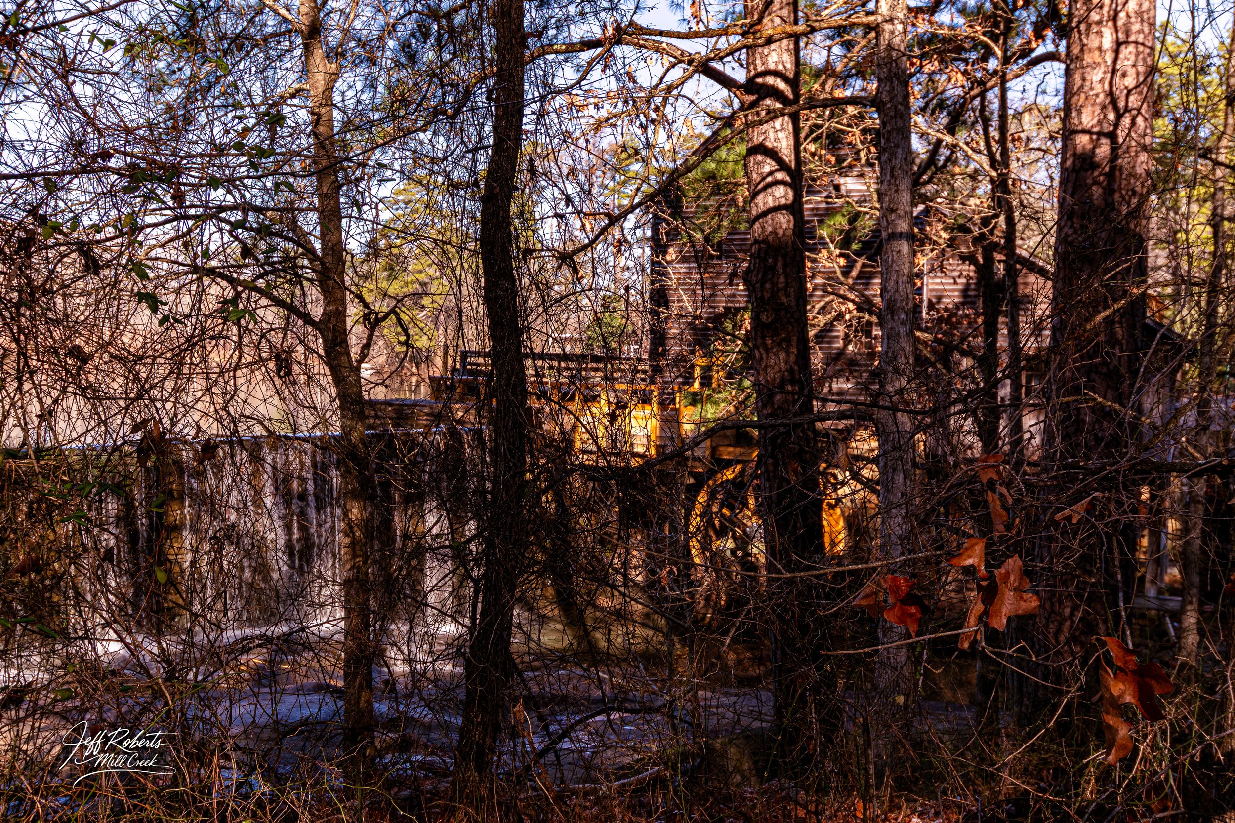 A rustic wooden watermill surrounded by trees, with water flowing over a small dam in the foreground. The scene appears to be in a wooded area with autumn leaves and natural sunlight.