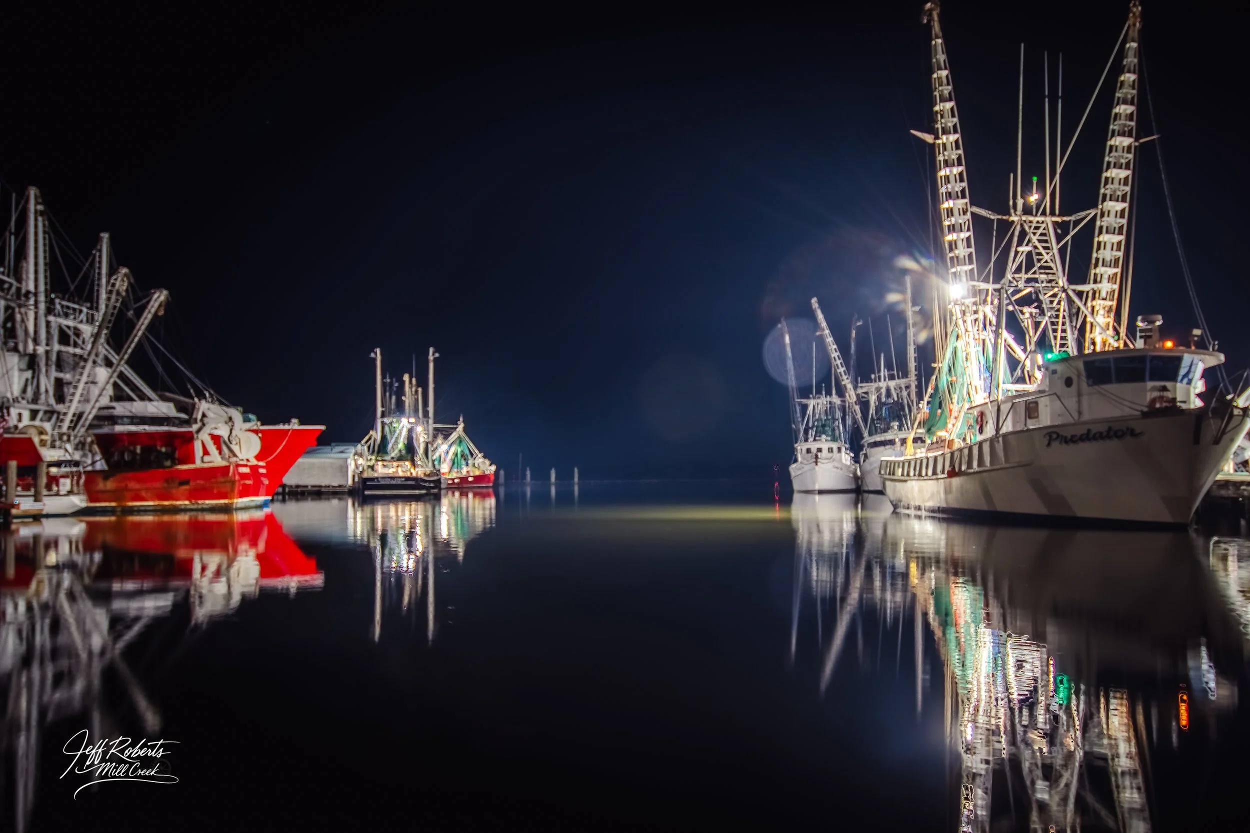 Nighttime view of docked fishing boats with their reflections on calm water, illuminated by boat lights.