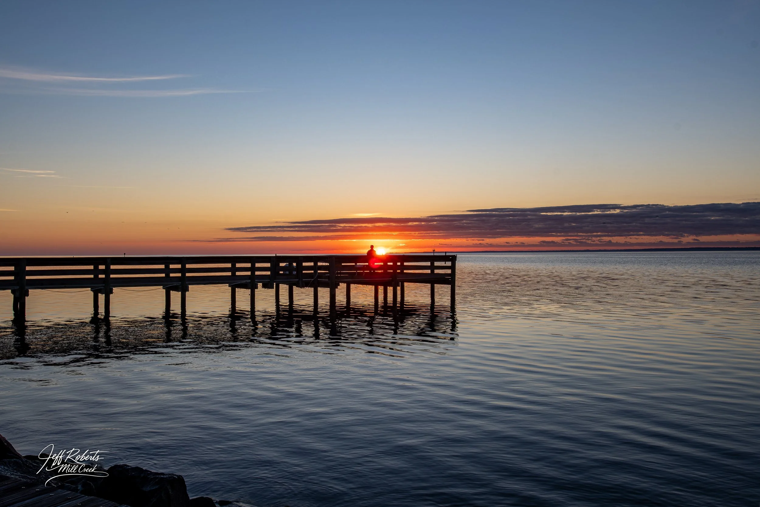 Sunset over a calm body of water with a wooden pier extending into the water, silhouette of a person sitting on the pier, and clouds on the horizon.