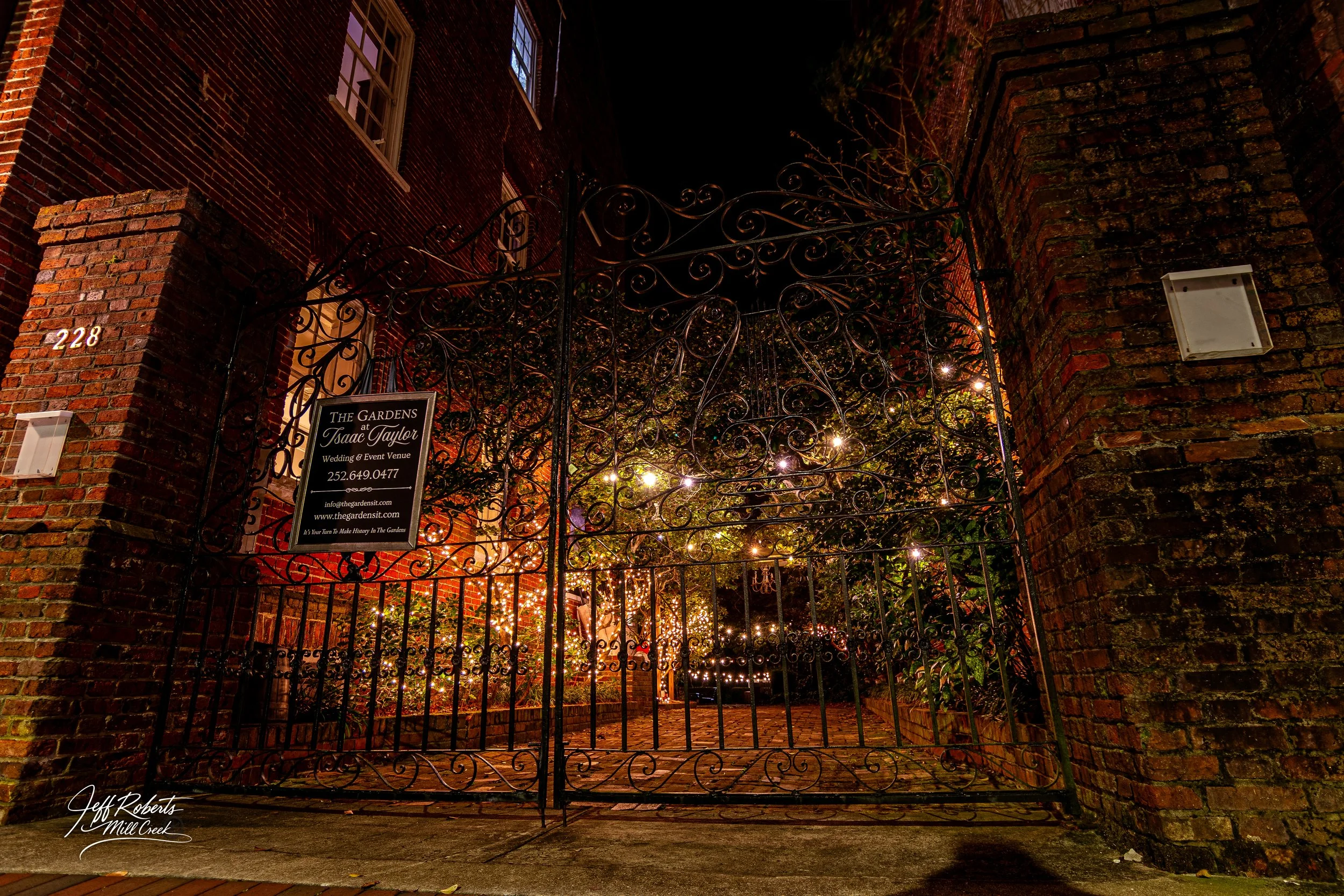 Night view of an ornate black wrought iron gate leading into a garden with fairy lights, brick building on the left, and brick wall on the right. Signboard on gate reads 'The Gardens at Isaac Taylor Wedding & Event Venue' with contact info.