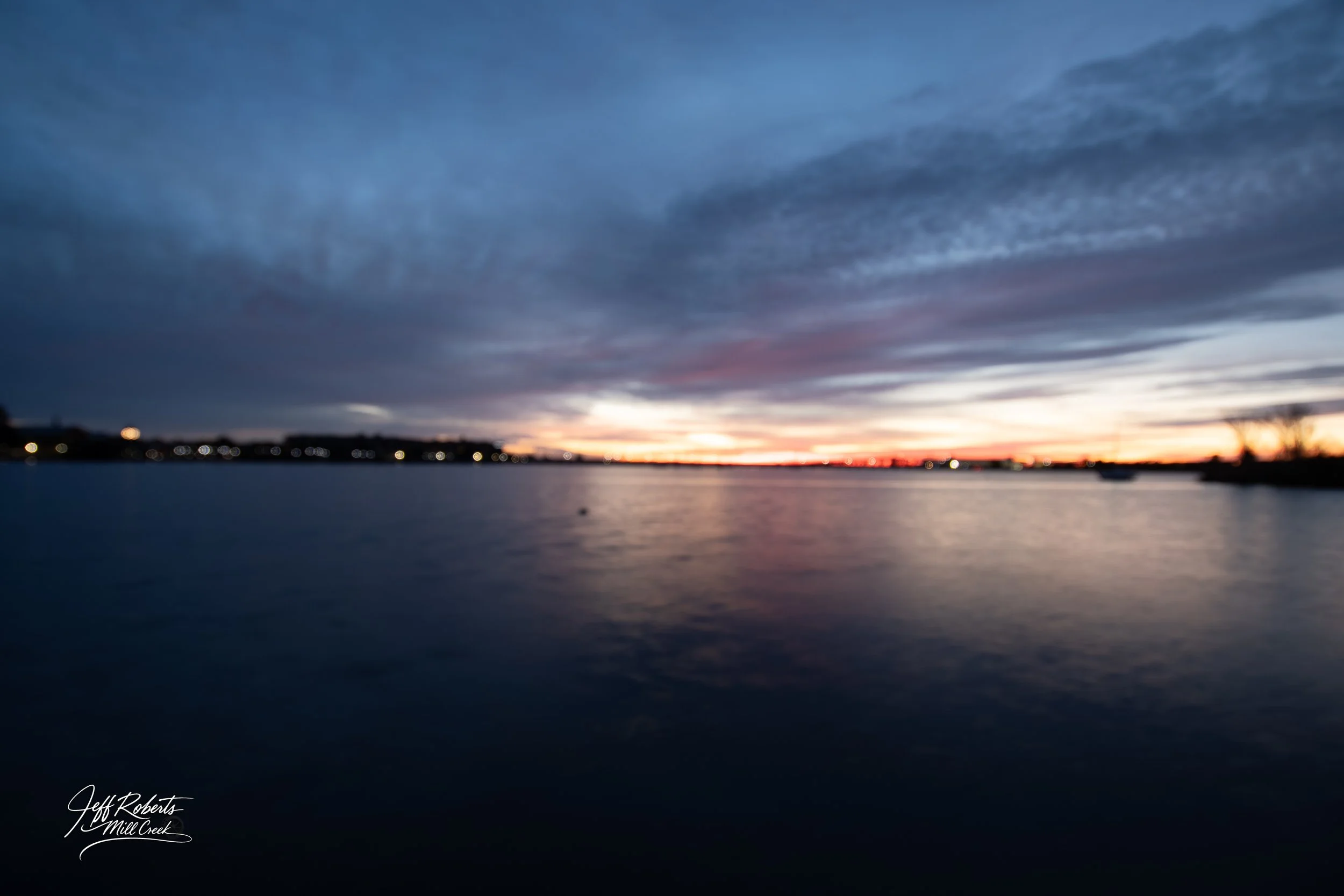 Sunset over a calm body of water, with dark clouds in the sky and distant city lights on the horizon.