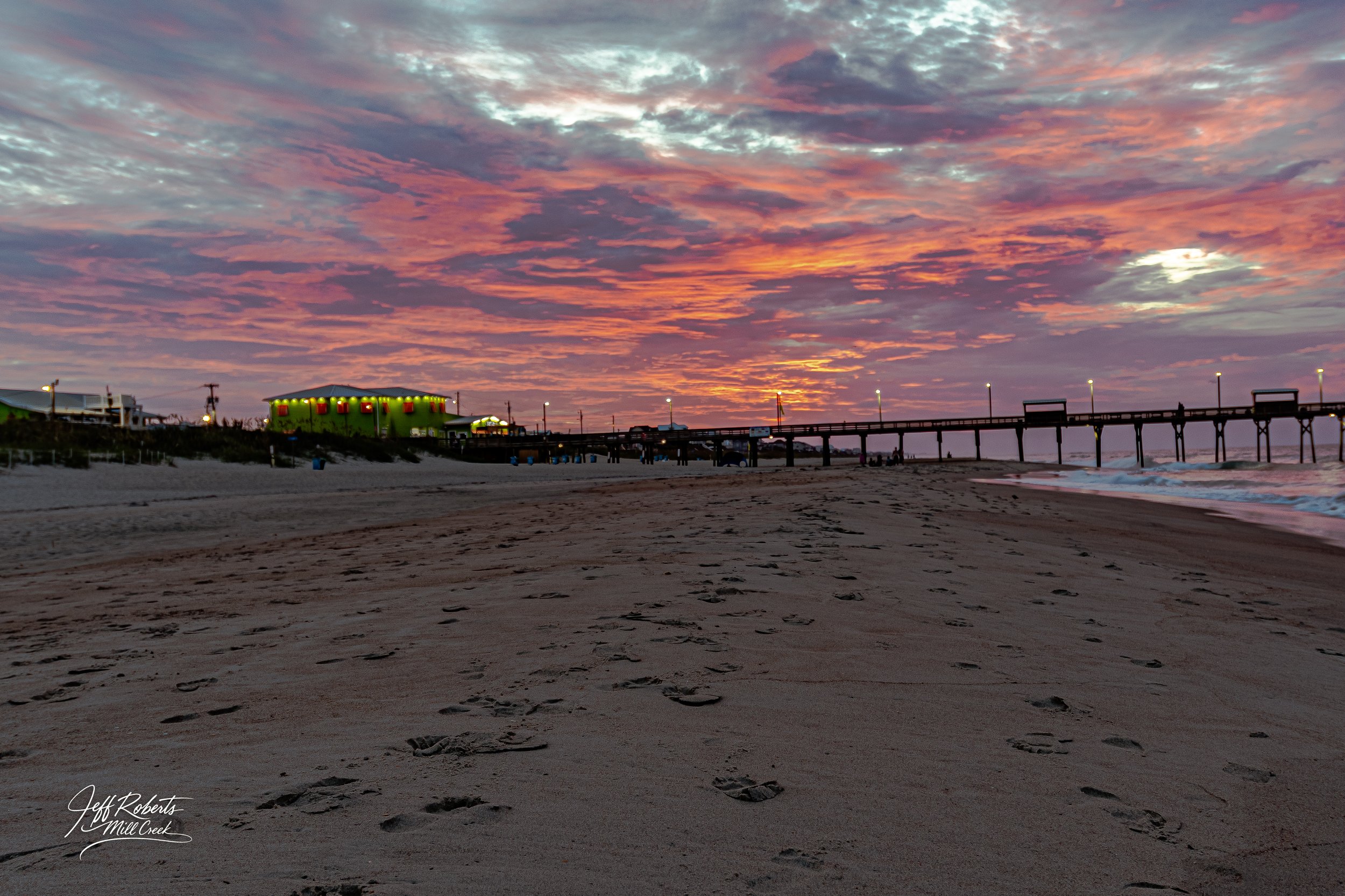Sunset over a sandy beach with footprints in the sand, a pier extending into the water, and a brightly lit green building in the background.