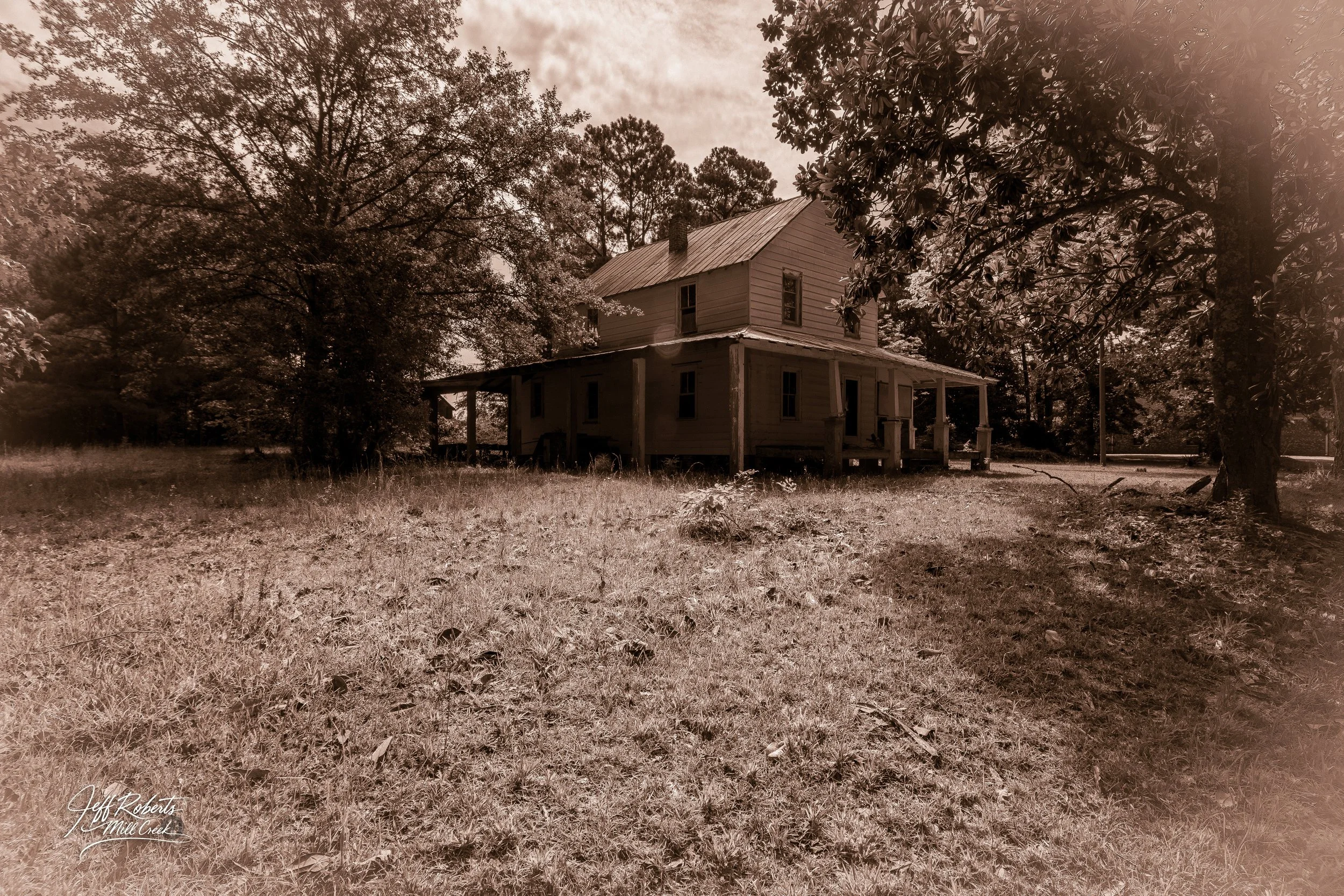 An old, two-story wooden house with a porch, surrounded by trees and grass in a rural setting, with sepia tone applied.