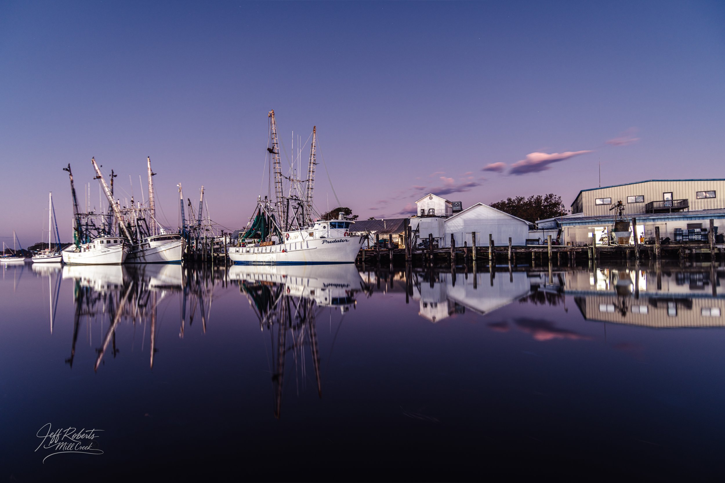 Boats docked at a marina during twilight, with buildings and trees in the background and their reflections mirrored in the calm water.