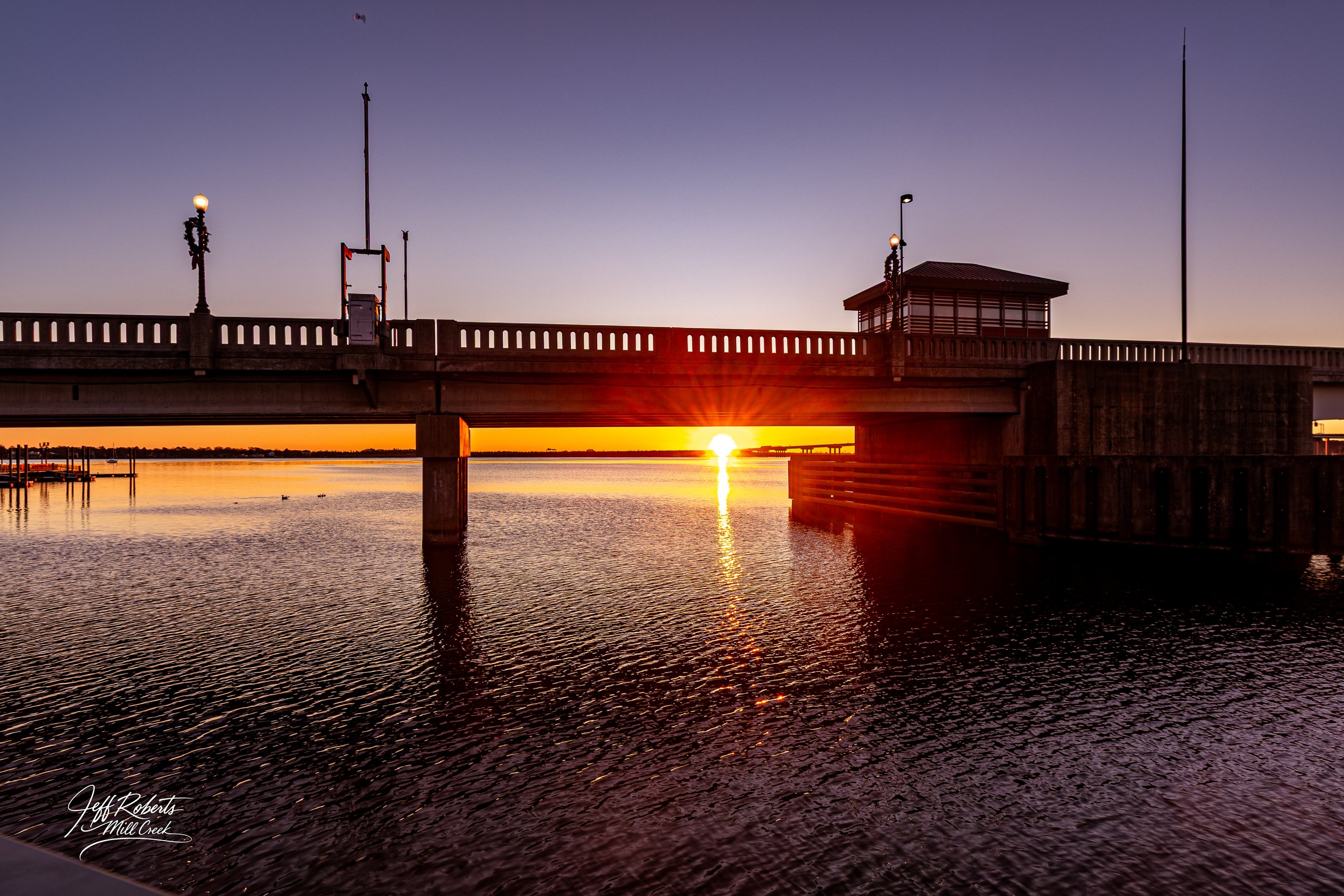 Sunset over a river with a bridge and a lamppost decorated with a Christmas wreath.