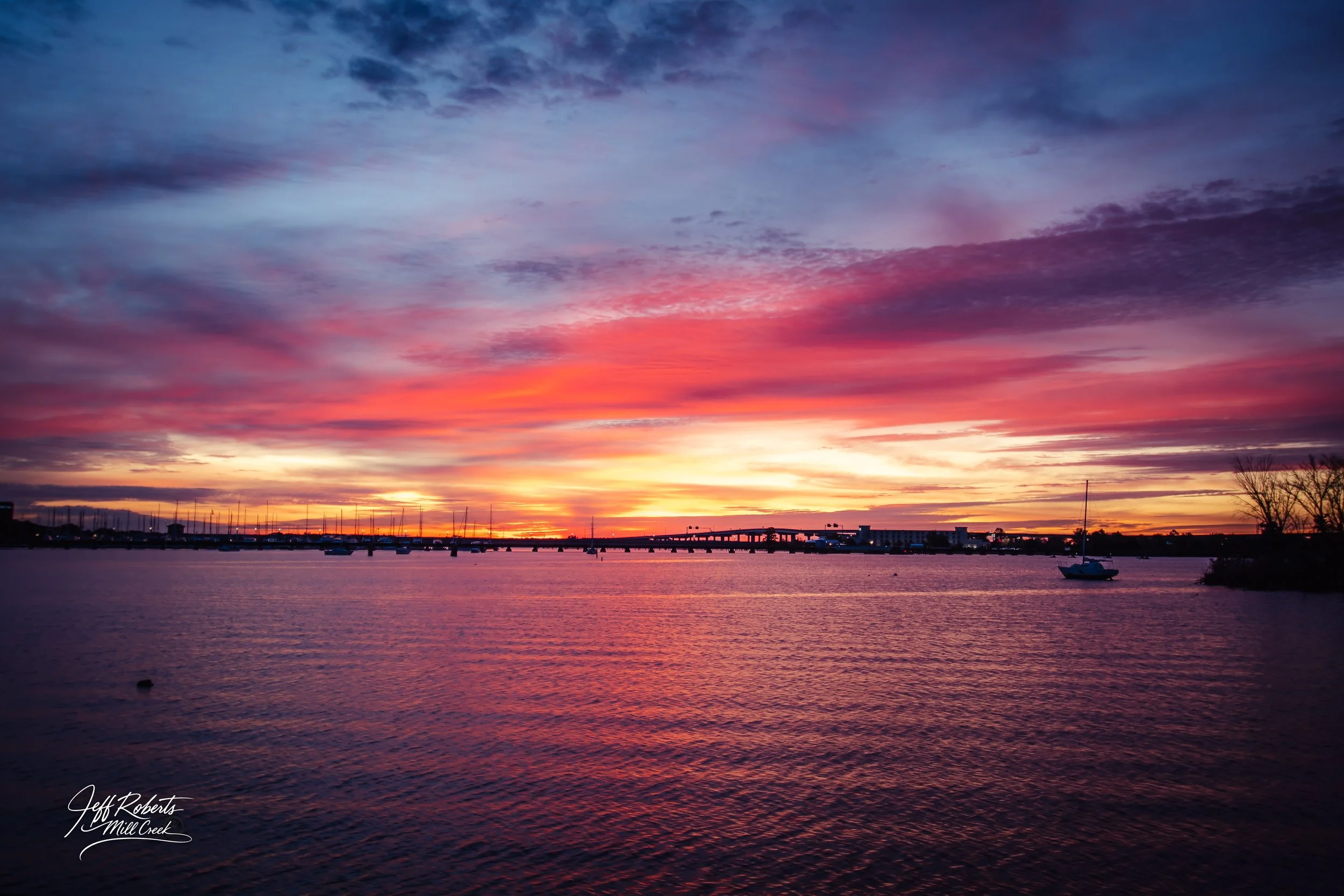 A colorful sunset over a bodies of water with boats and a bridge in the distance, with vibrant pink, purple, and orange hues in the sky.
