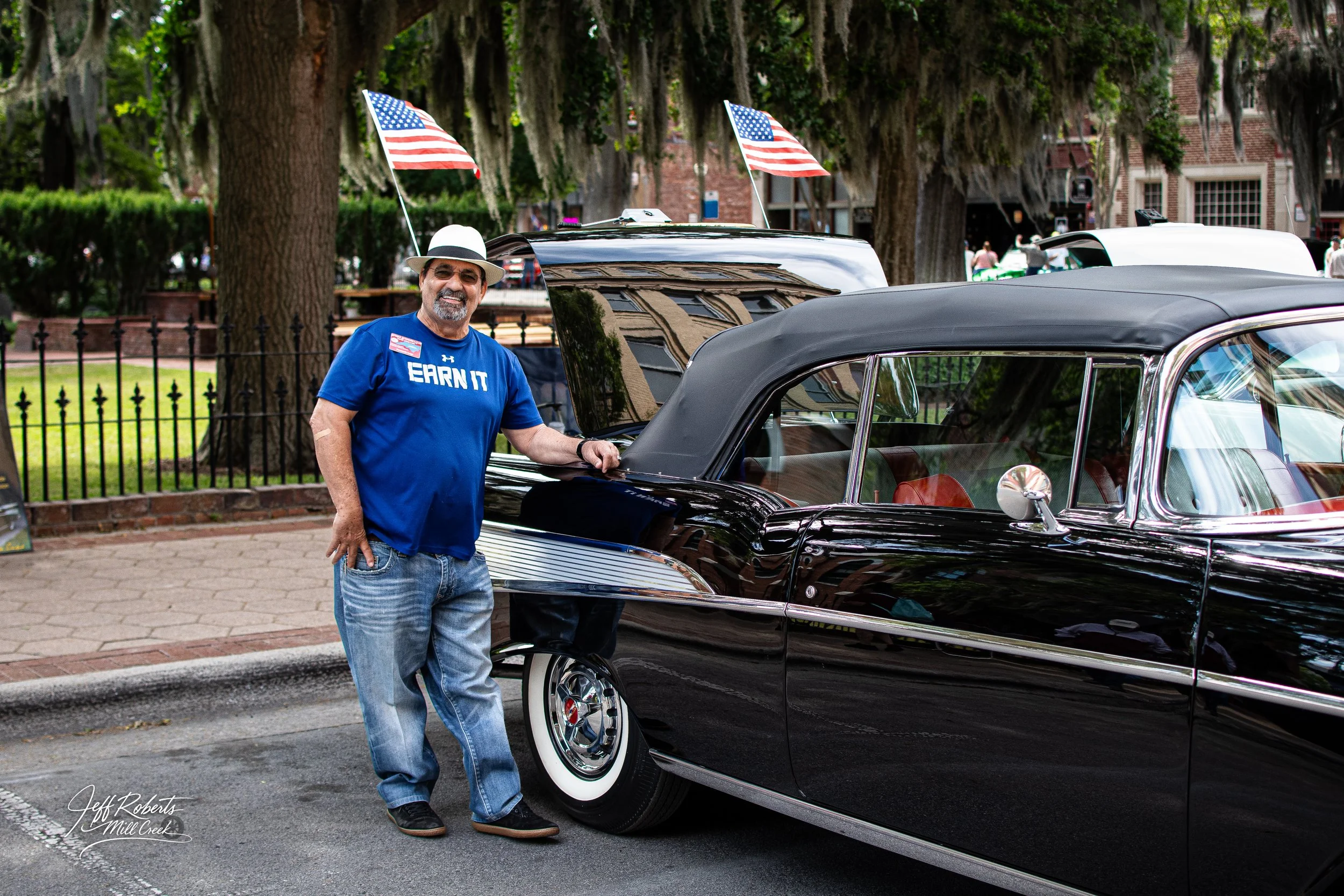 A man in a blue T-shirt, jeans, and a fedora hat standing next to a classic black car with a convertible top, at an outdoor car show with American flags and trees in the background.