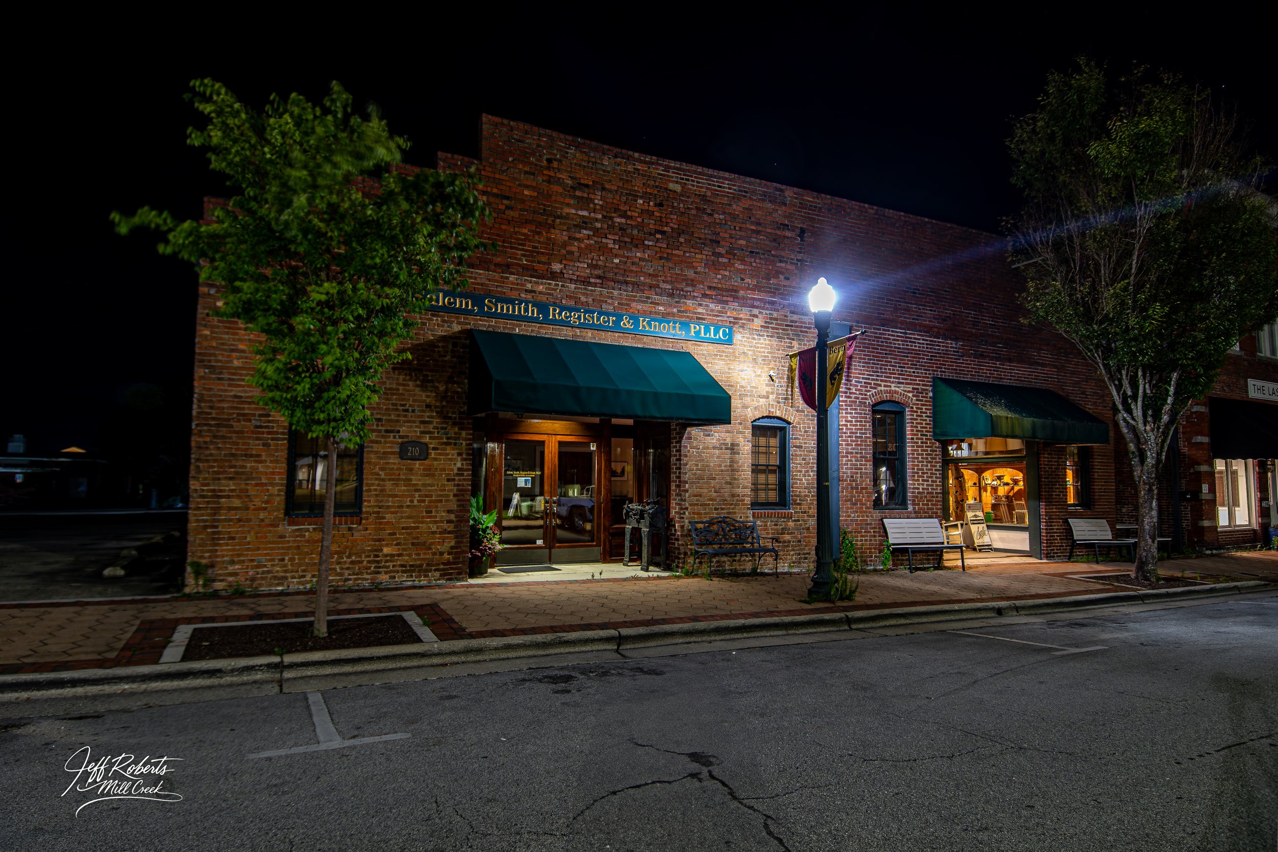 Nighttime view of a brick building with illuminated signs, green awnings, benches, and trees on the sidewalk, streetlights, and interior lights visible in the windows.