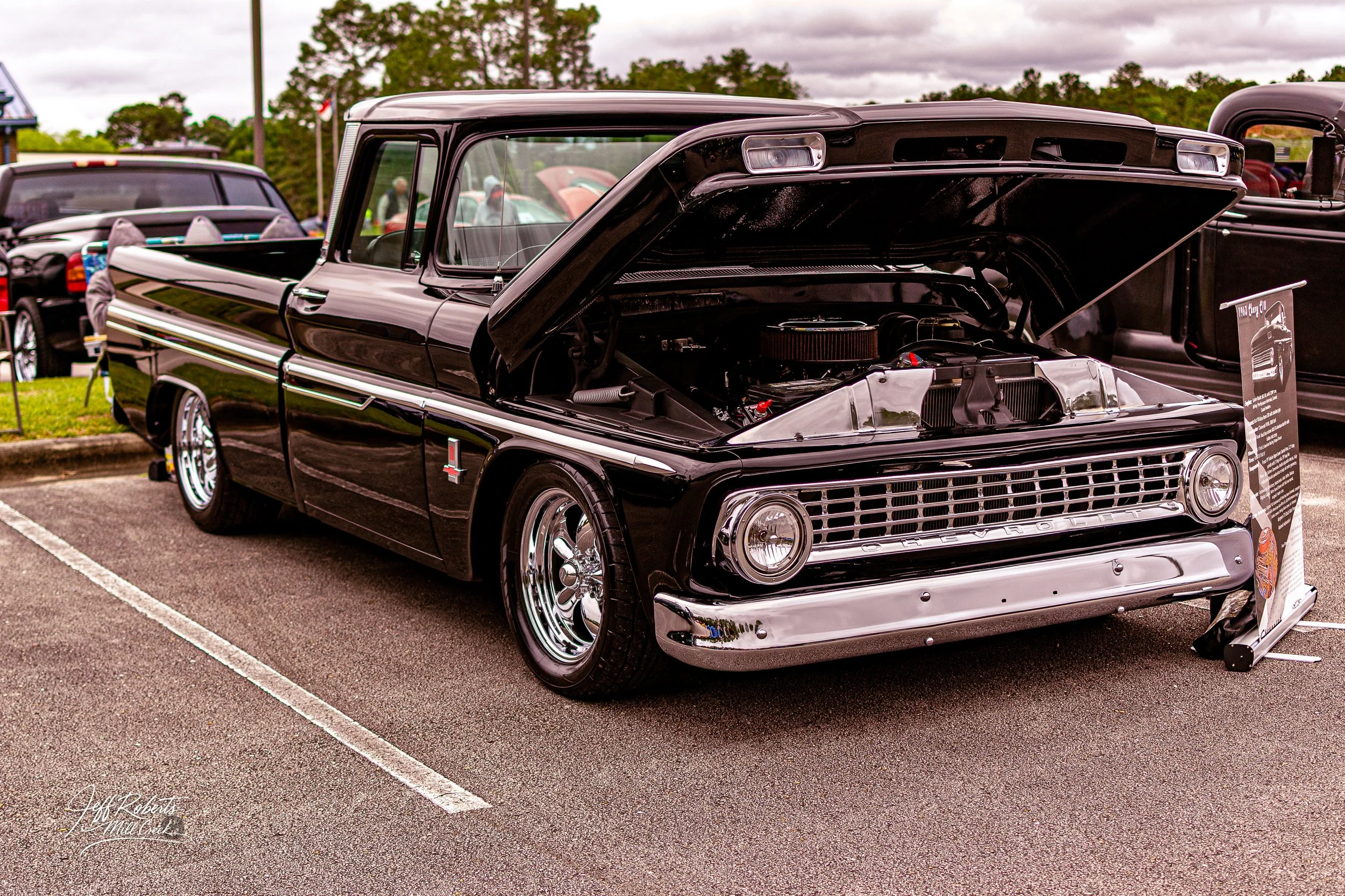 Black vintage Chevrolet pickup truck with open hood, showing engine, parked at a car show with other cars in background