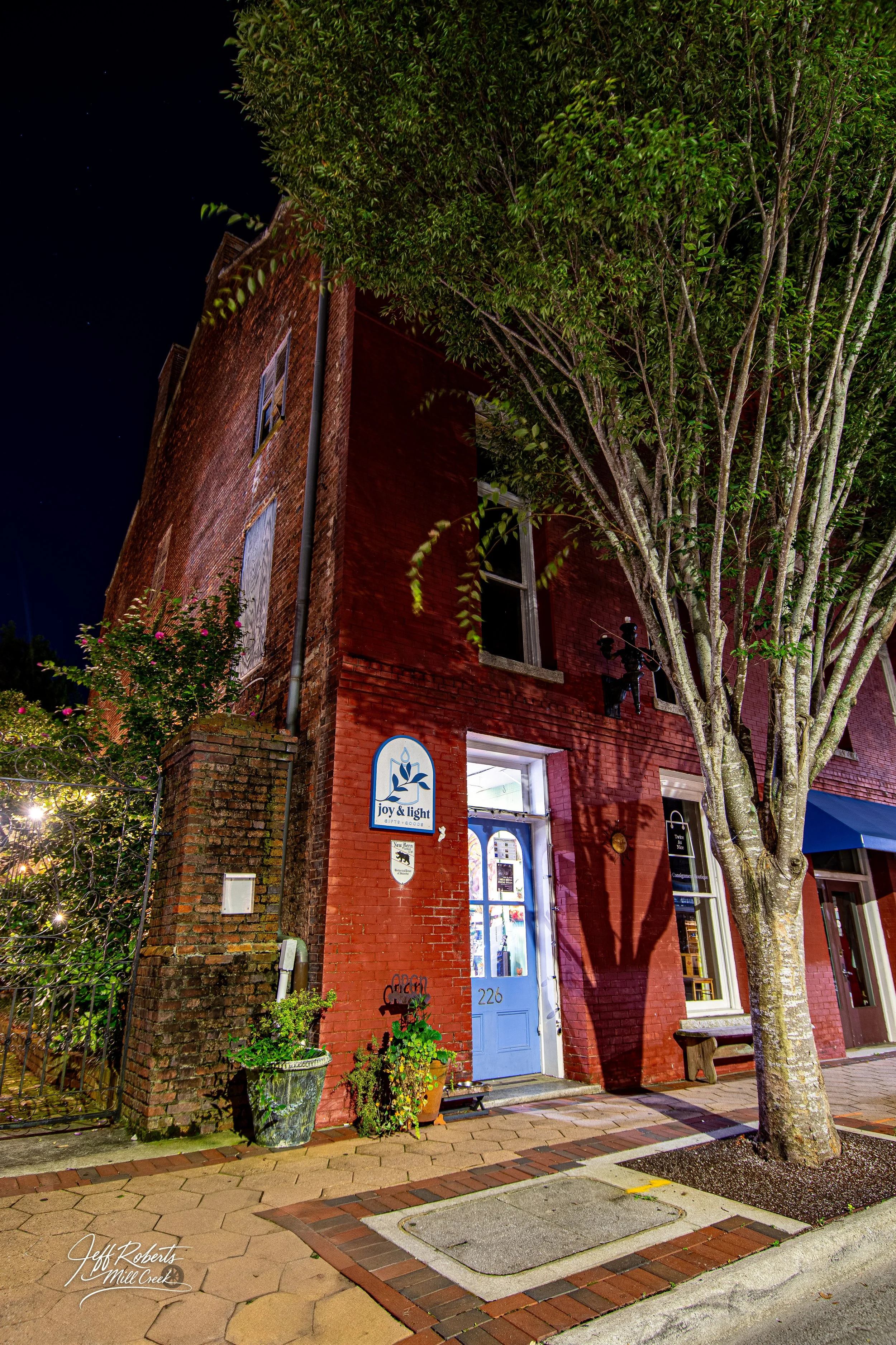 A brick building at night with a blue door, illuminated window displays, a large tree, and a street sign reading "joy & light." There are potted plants and a bench outside, and the scene is lit by streetlights.