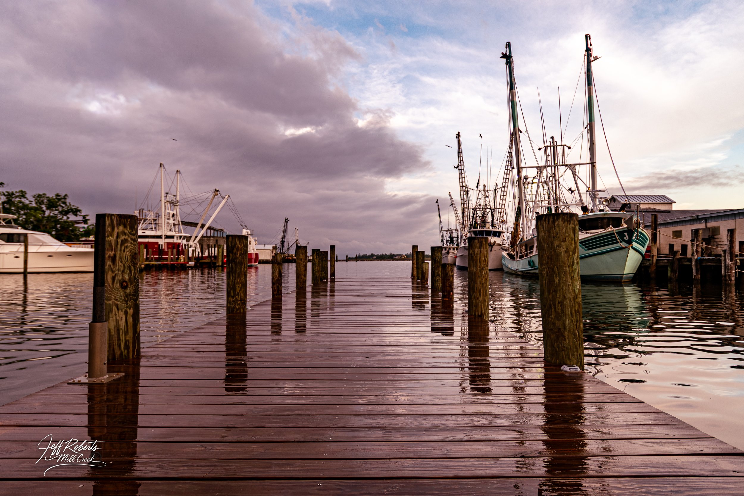 A wooden dock extending into a harbor with boats docked on both sides under a cloudy sky at sunset.