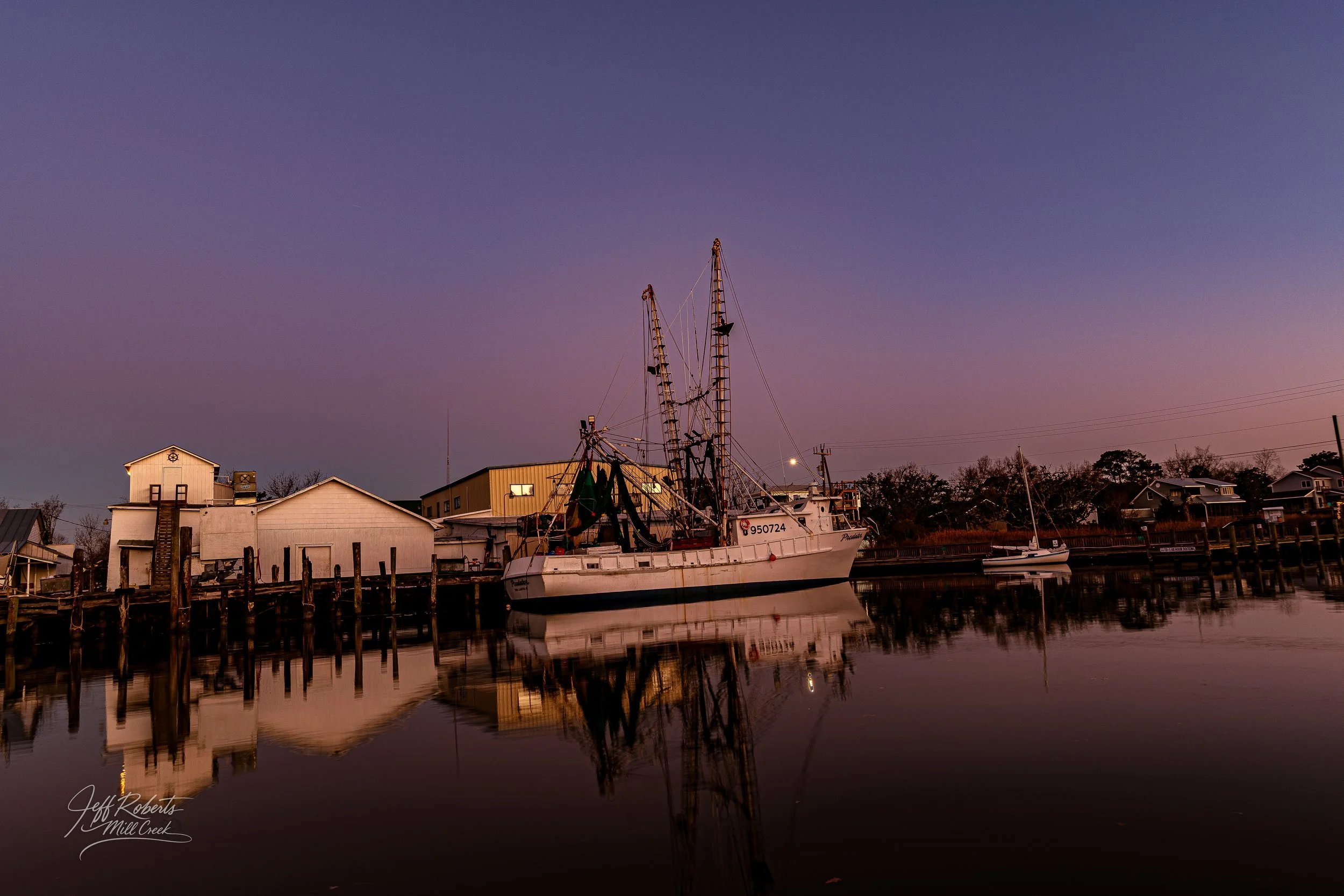 A boat docked at a pier during twilight with calm water reflecting the boat and surrounding buildings.