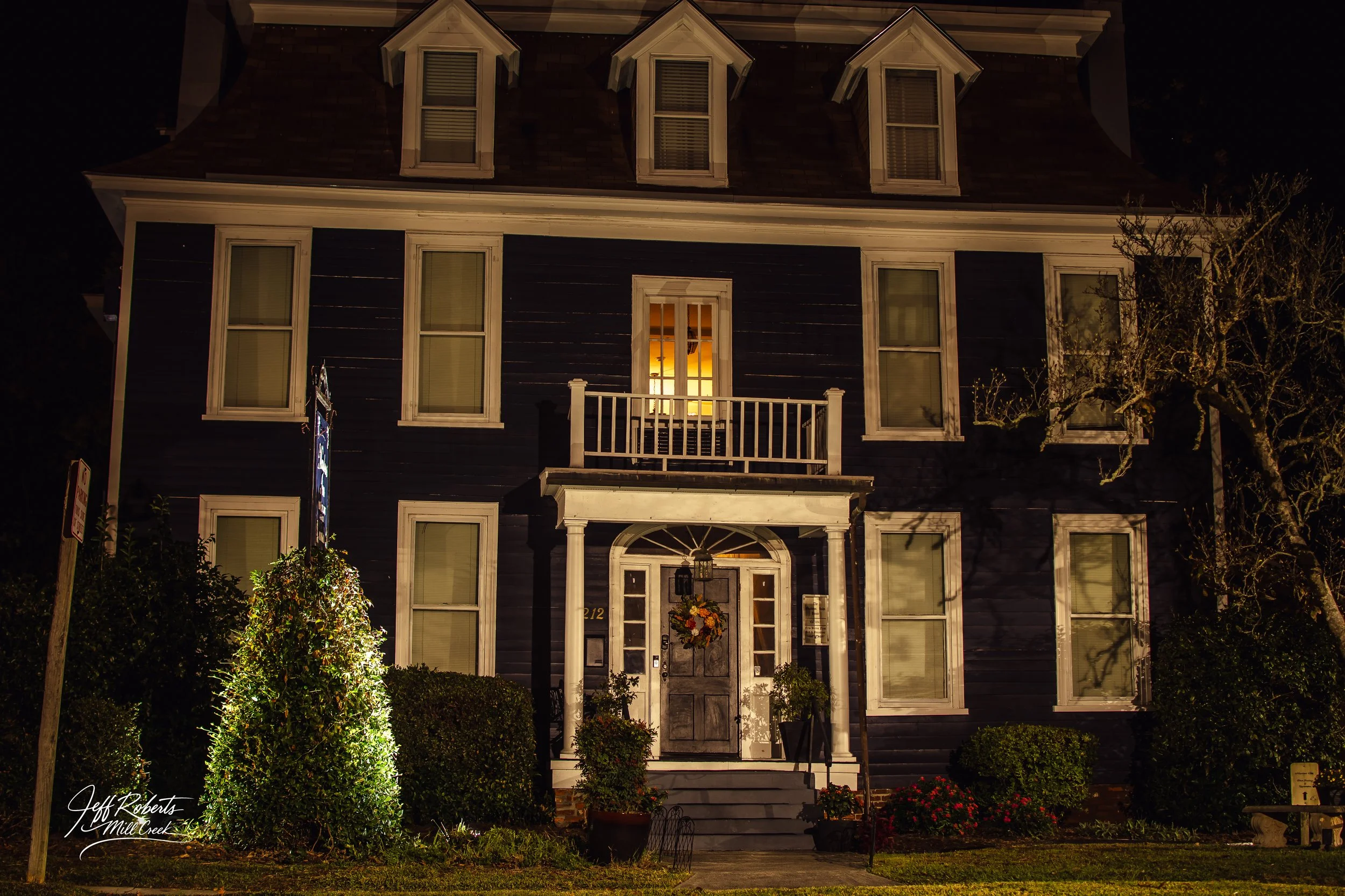 A dark blue, three-story house with white trim, illuminated by exterior lighting at night. The house has six large windows, a small balcony above the front door, and a decorated front porch with stairs. Flowers and bushes are in the front yard, and a