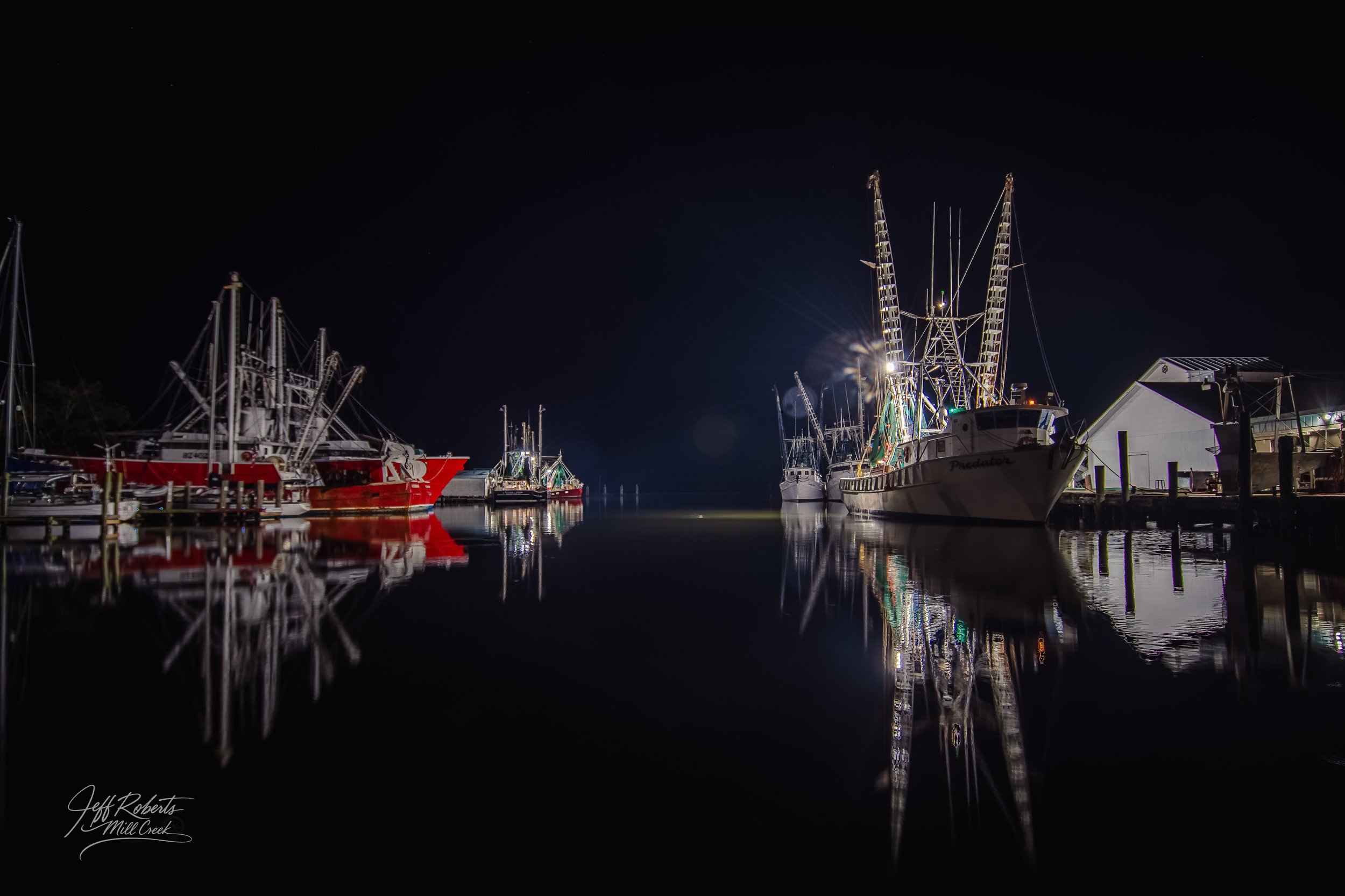 Nighttime scene of boats docked at a marina, with reflections visible in the calm water, and a dark sky in the background.