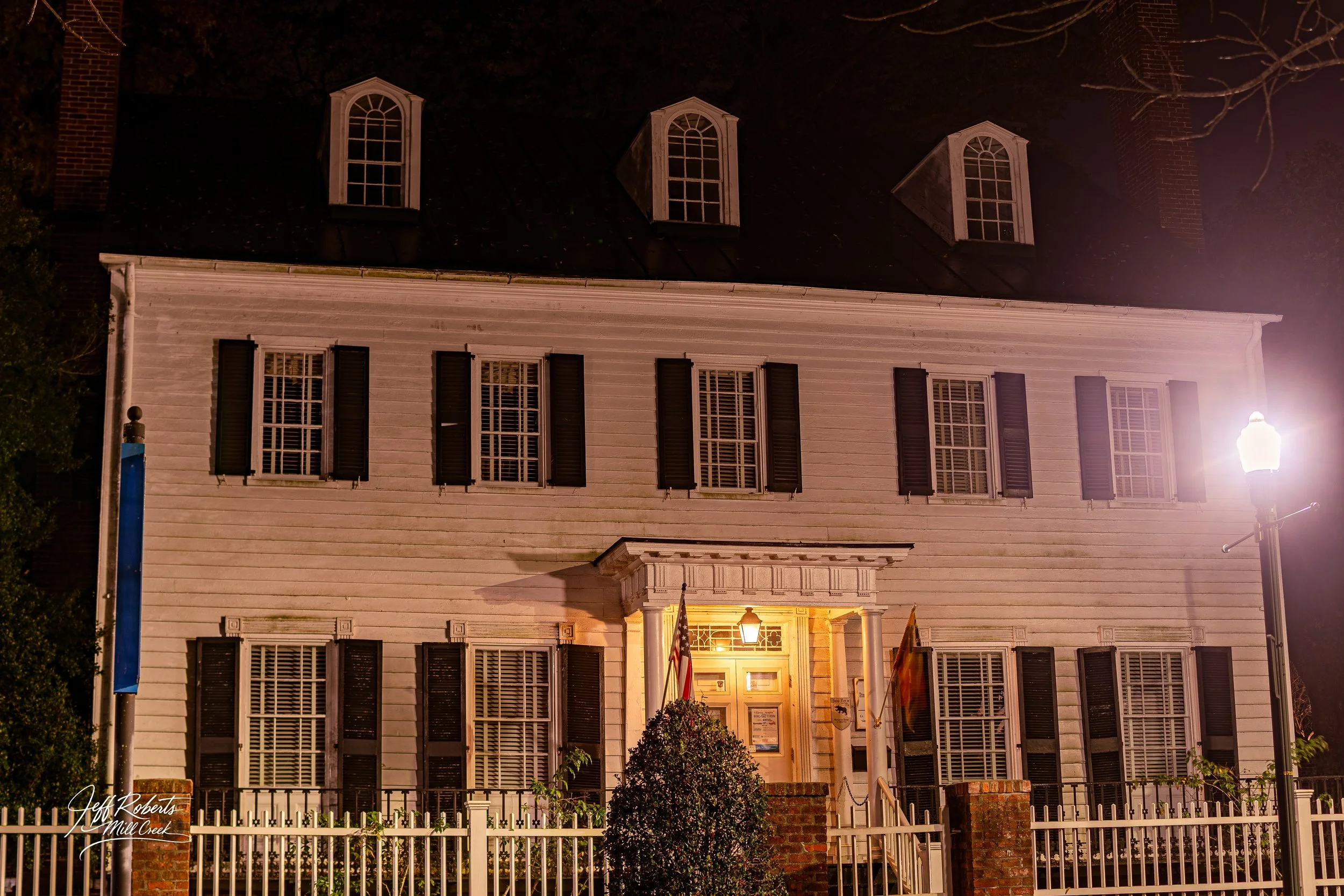 A three-story white wooden building with black shutters and a covered front porch illuminated by a hanging front lantern, surrounded by a white picket fence, at night.