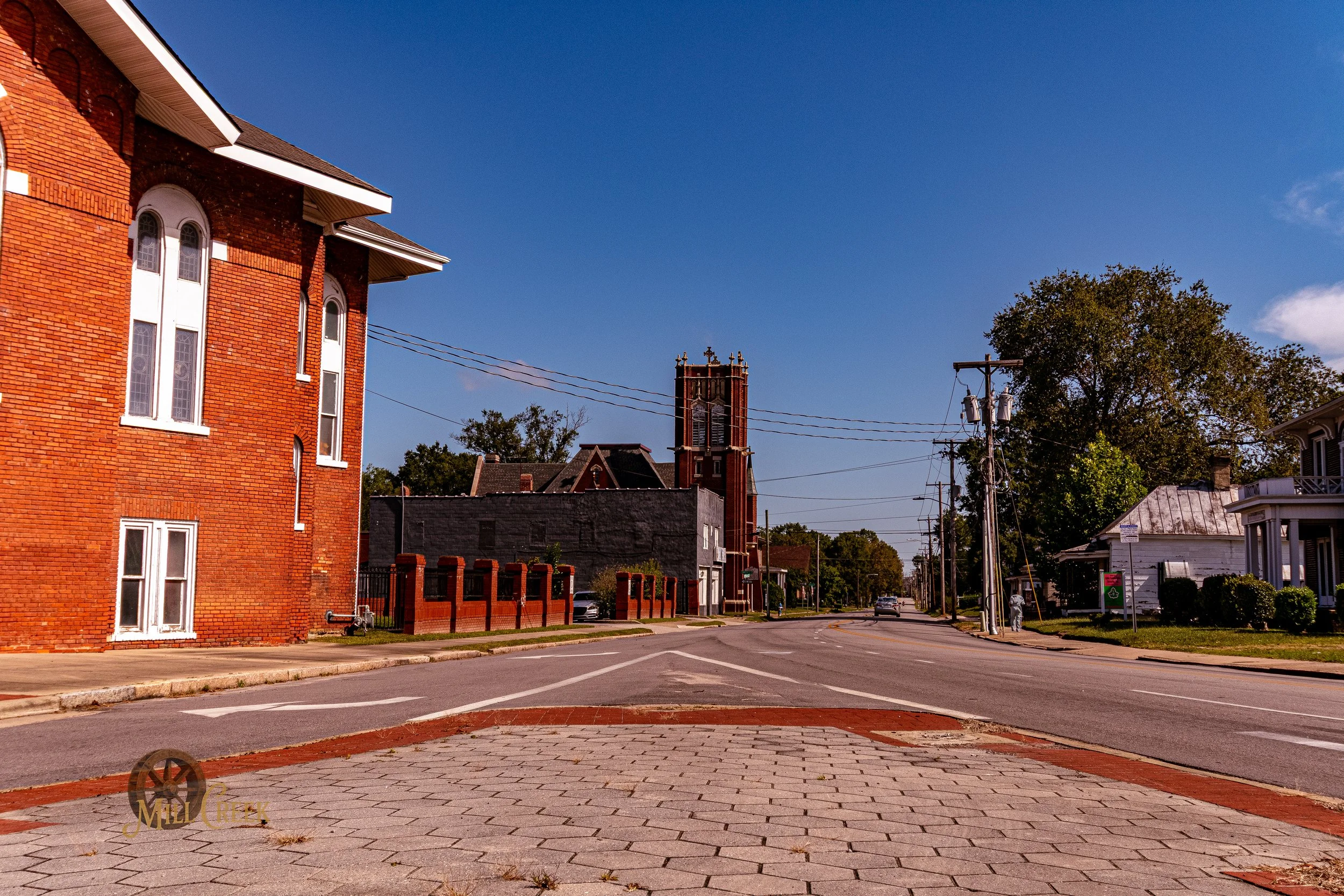 A small town street scene featuring a prominent red brick church with a tall steeple, surrounded by residential houses and a bright blue sky.