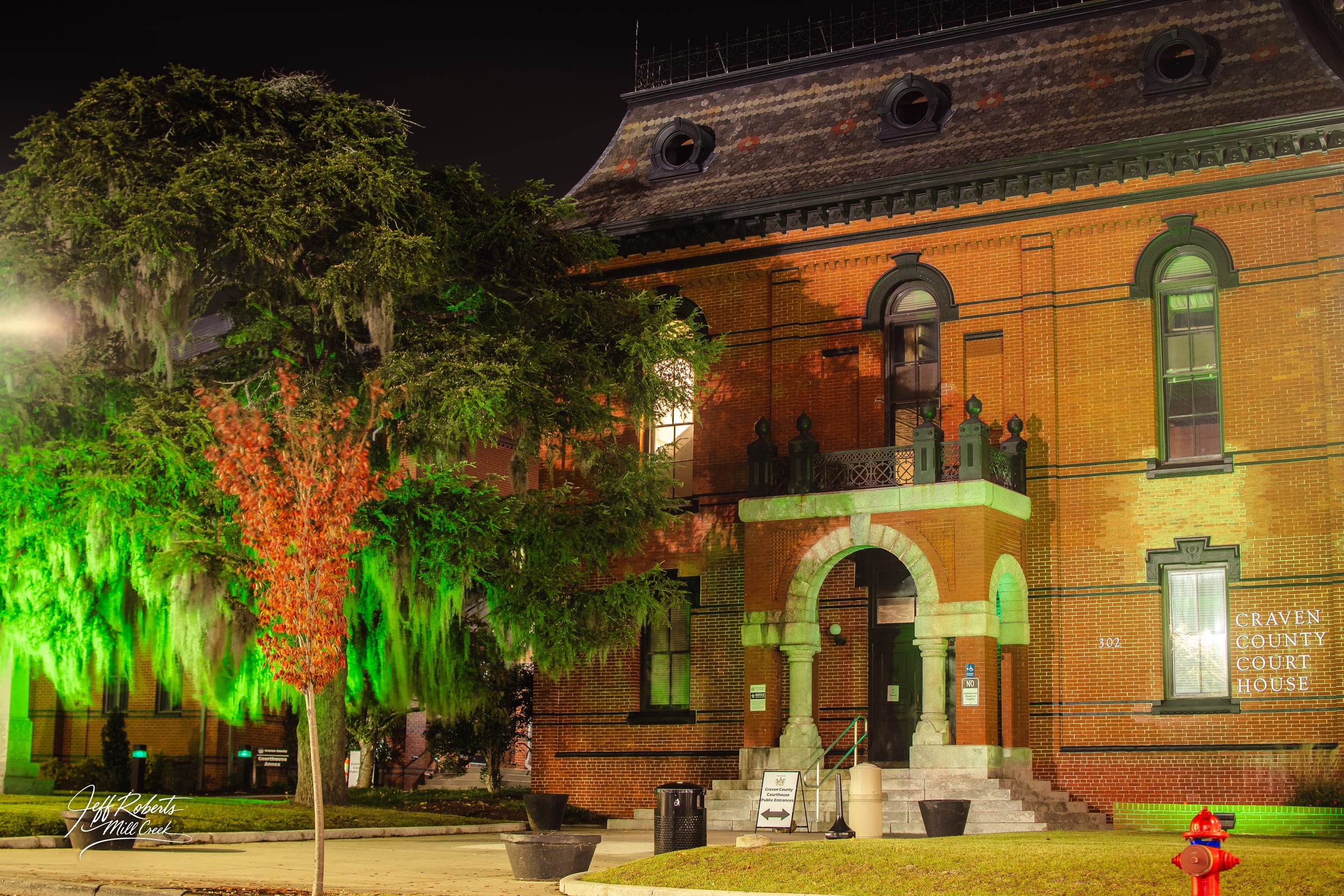 Nighttime view of the Craven County Courthouse in North Carolina, a historic red brick building with arched windows and a small balcony. The courthouse is illuminated with green and yellow lights, and there are trees with green and orange leaves in f