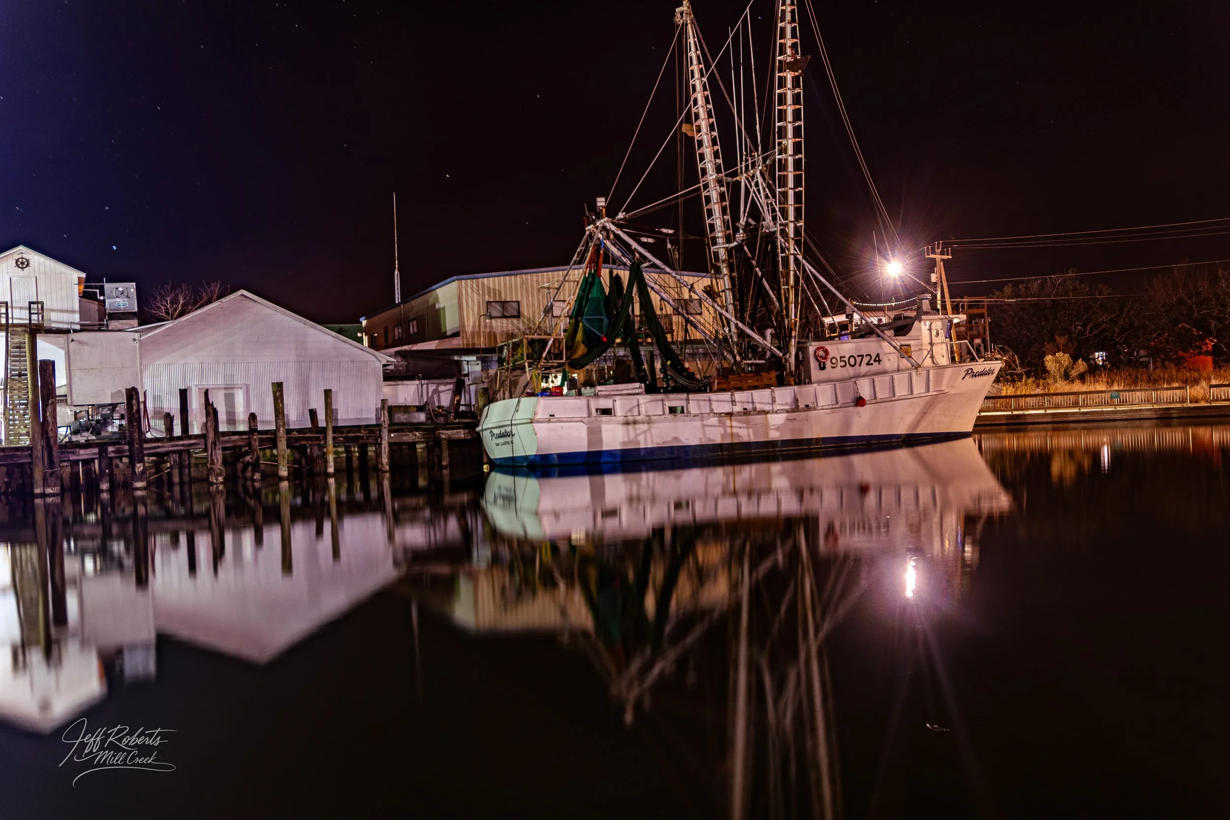 A docked fishing boat named 'Predator' reflecting on calm water at night, with buildings and power lines in the background, under a starry sky.