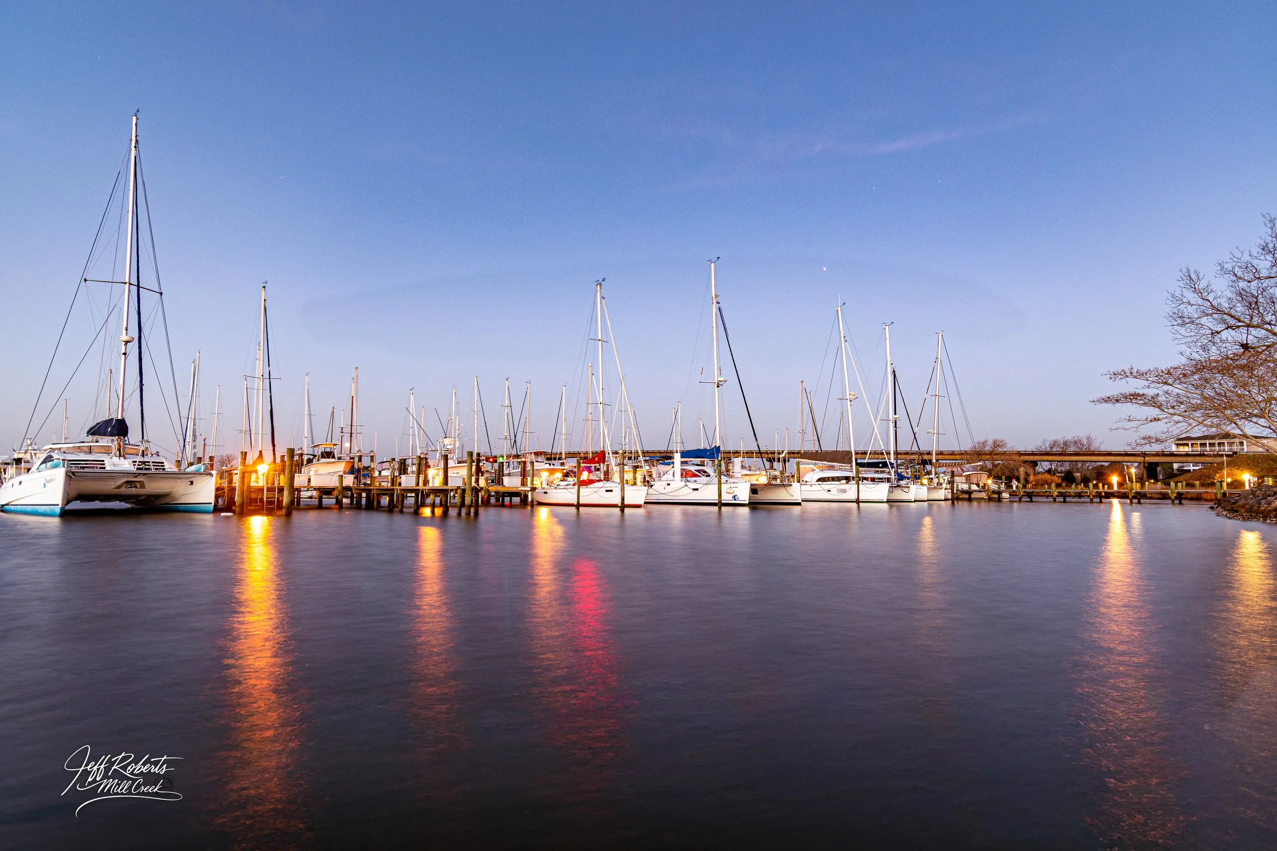 A marina with sailboats docked at the pier during dusk, with reflections of lights on calm water and a clear sky overhead.