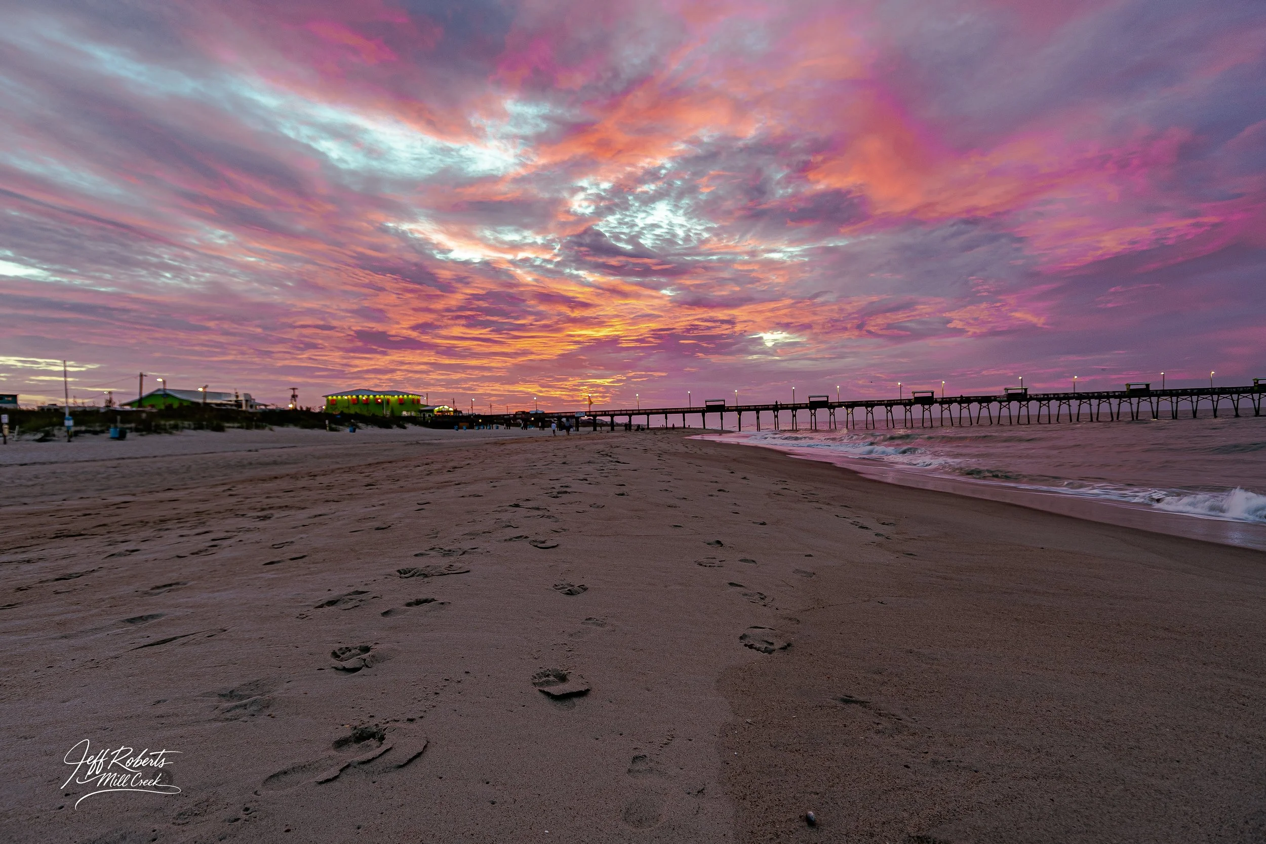 A sandy beach at sunset with colorful pink, purple, and orange clouds in the sky, footprints in the sand, a pier extending into the ocean, and a building with green lights in the distance.