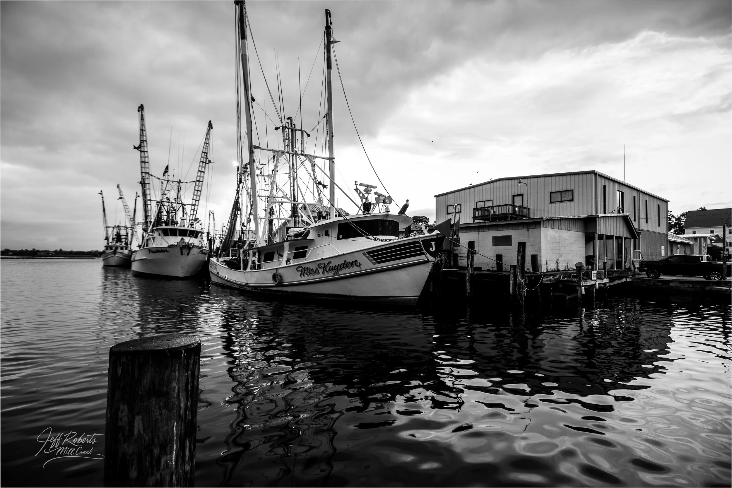 Black and white photo of boats docked at a marina next to a modern building, with water, reflections, and cloudy sky in the background.