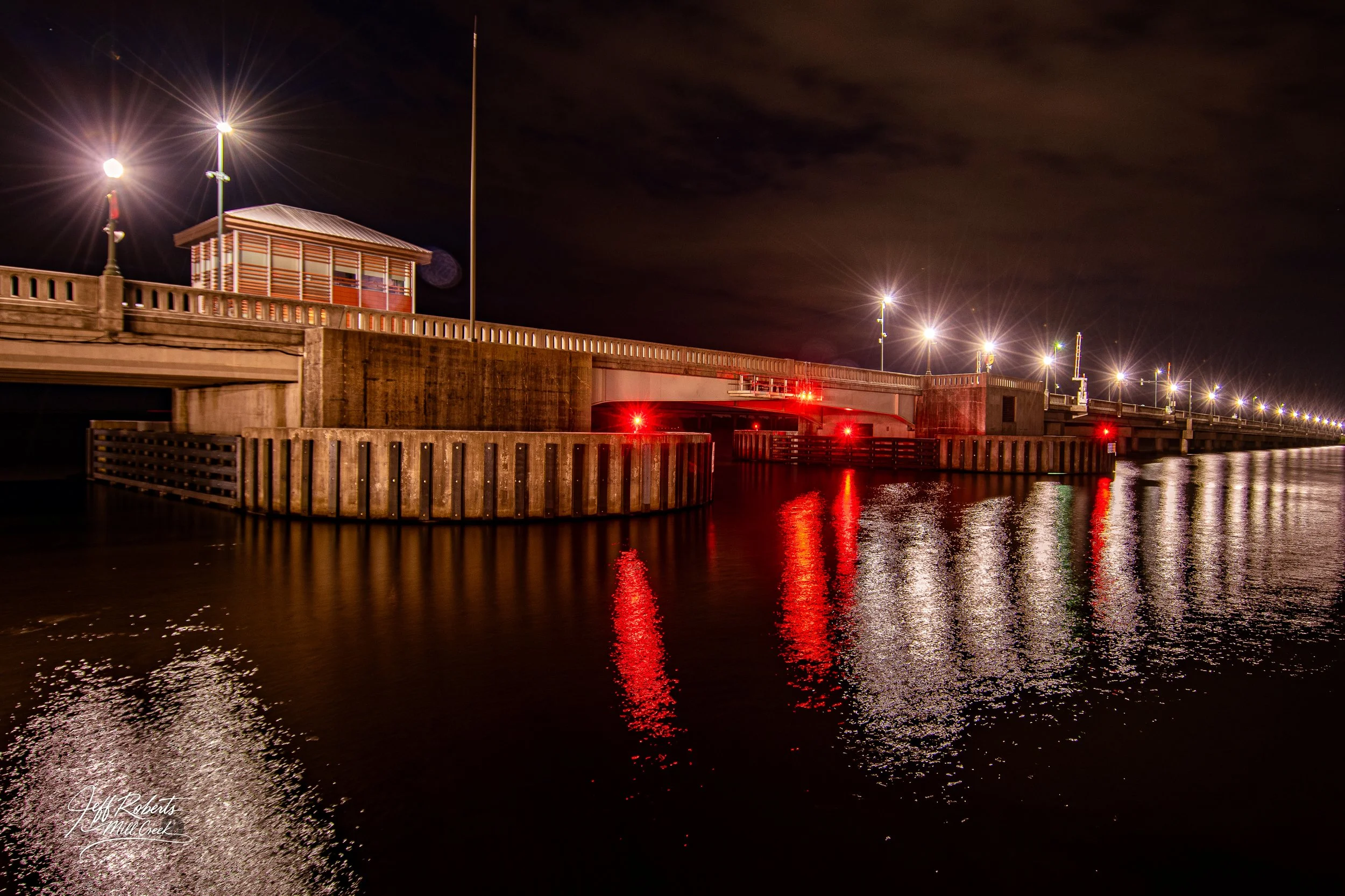 Night view of a lit bridge over water with reflections and a small structure on top, illuminated by streetlights.
