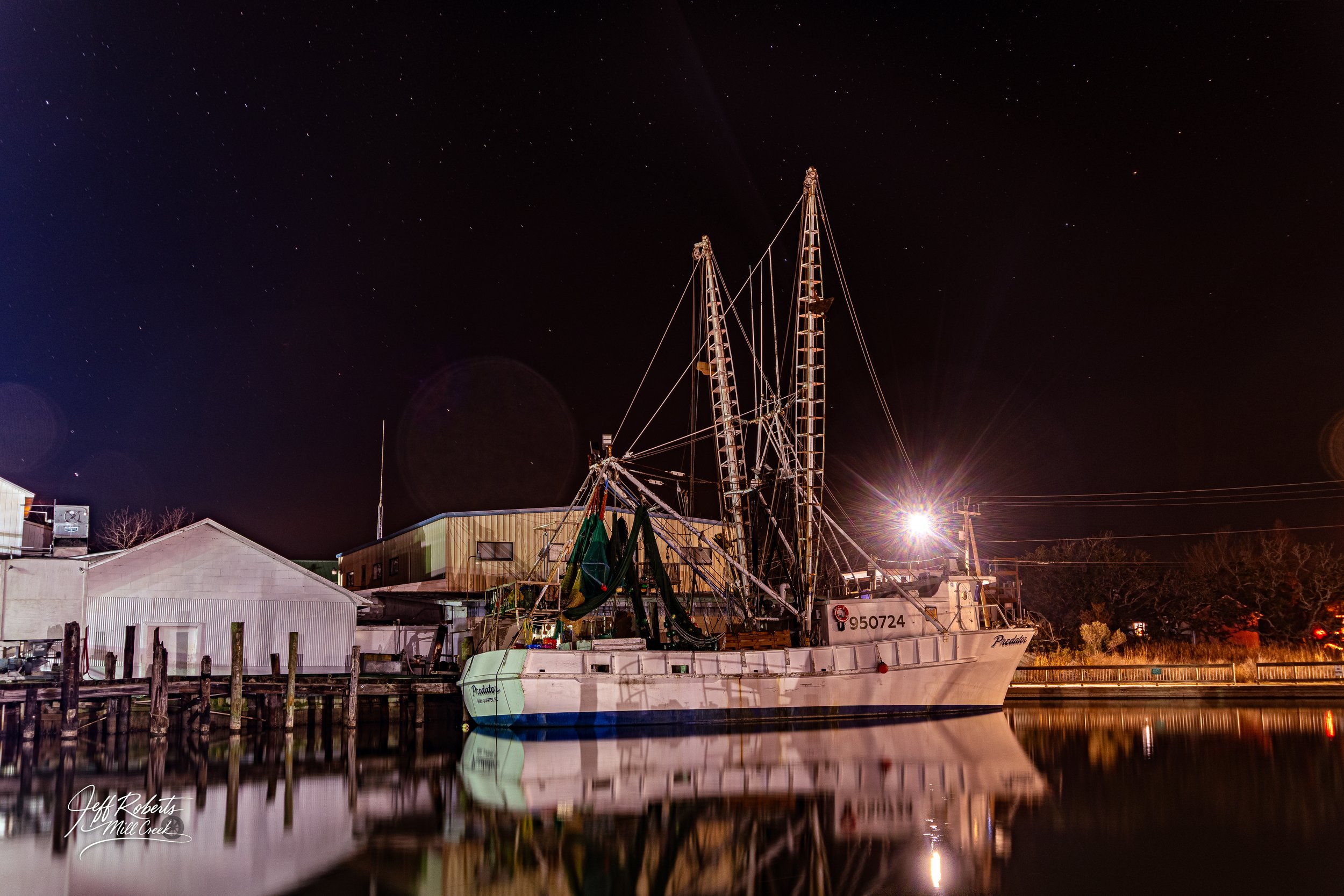 A fishing boat docked at night with a starry sky above, reflected in calm water, near buildings and trees with a bright light shining from the right side.