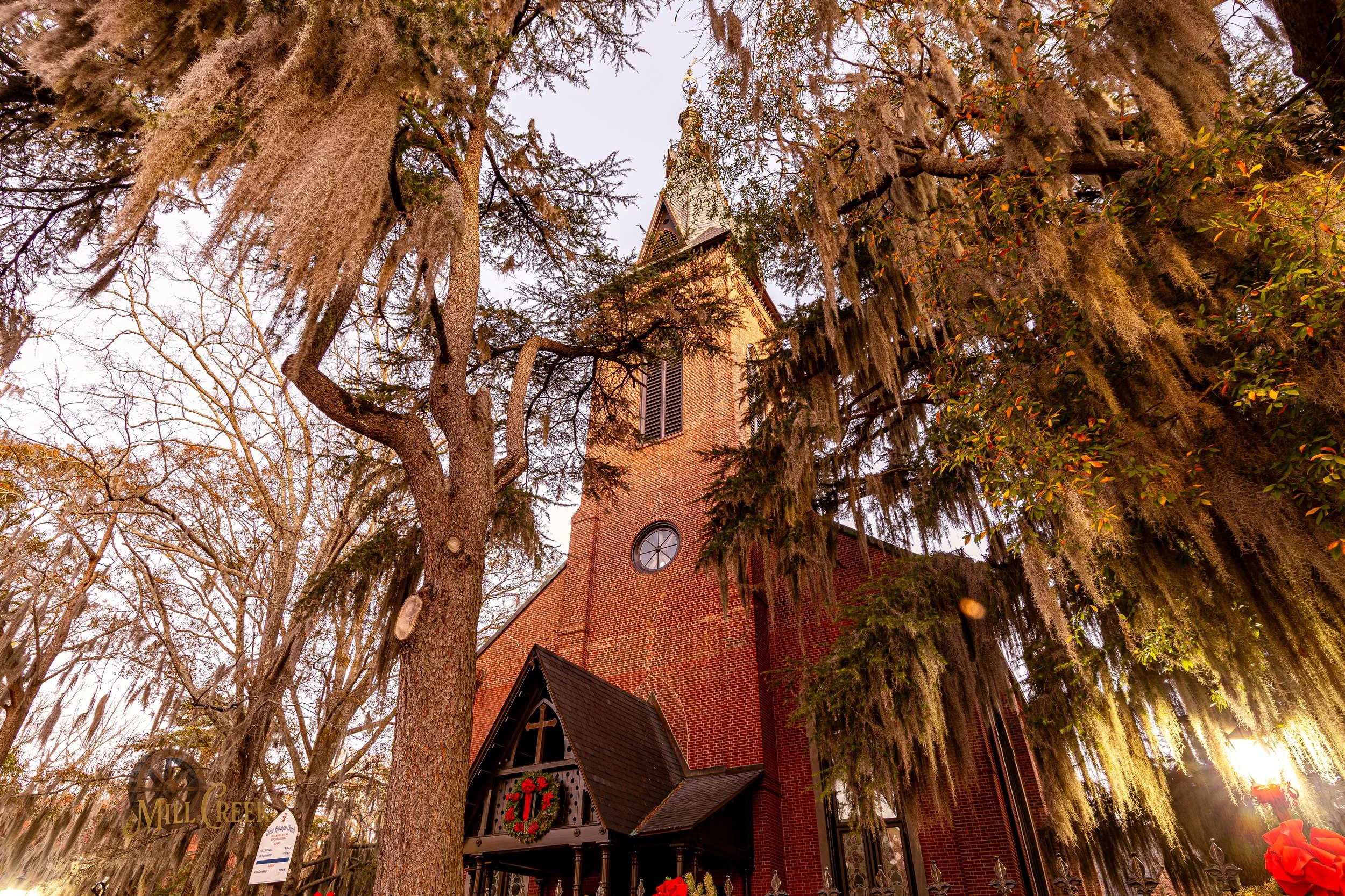 A brick church with a tall steeple surrounded by large trees with moss hanging from their branches, and decorated with a Christmas wreath on the entrance.