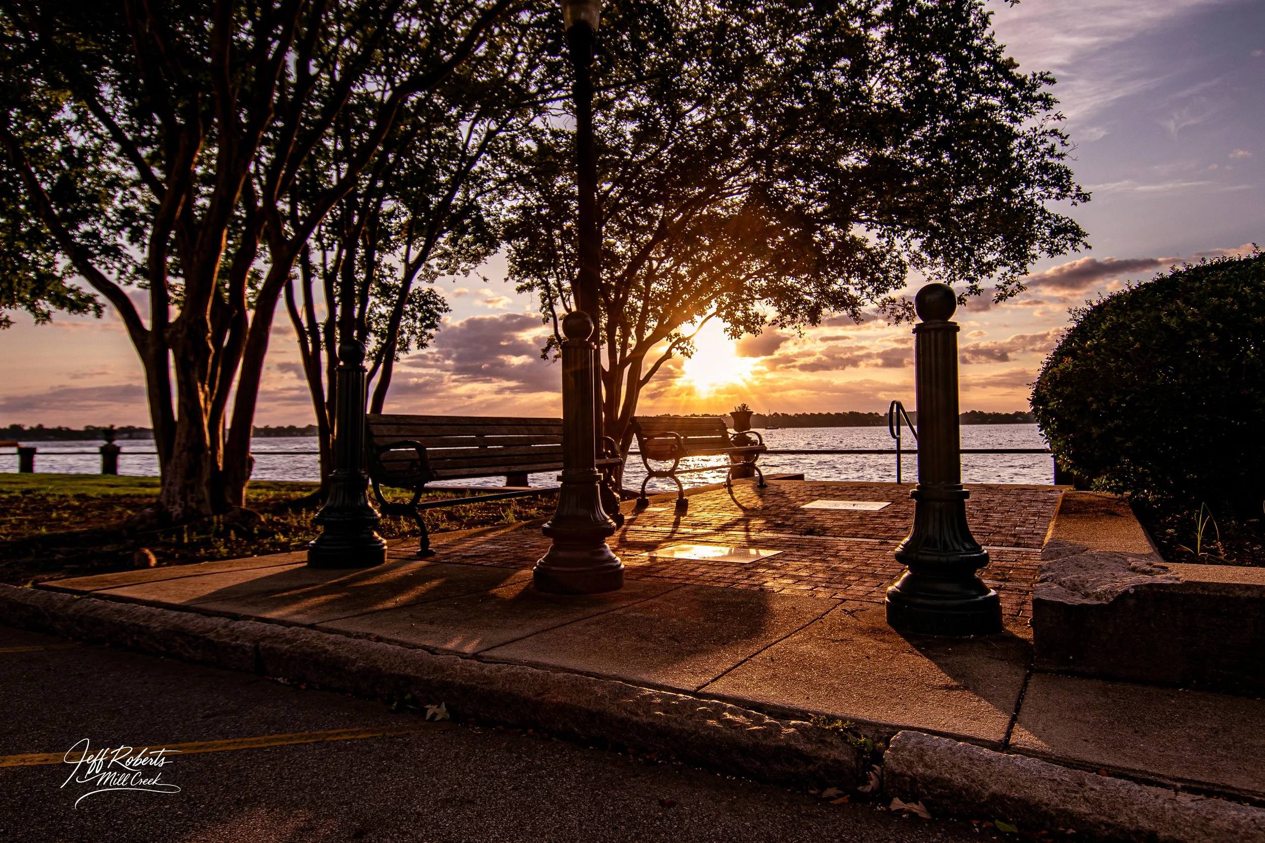 Sunset over a river viewed from a park with benches, trees, lamp posts, and a paved walkway.
