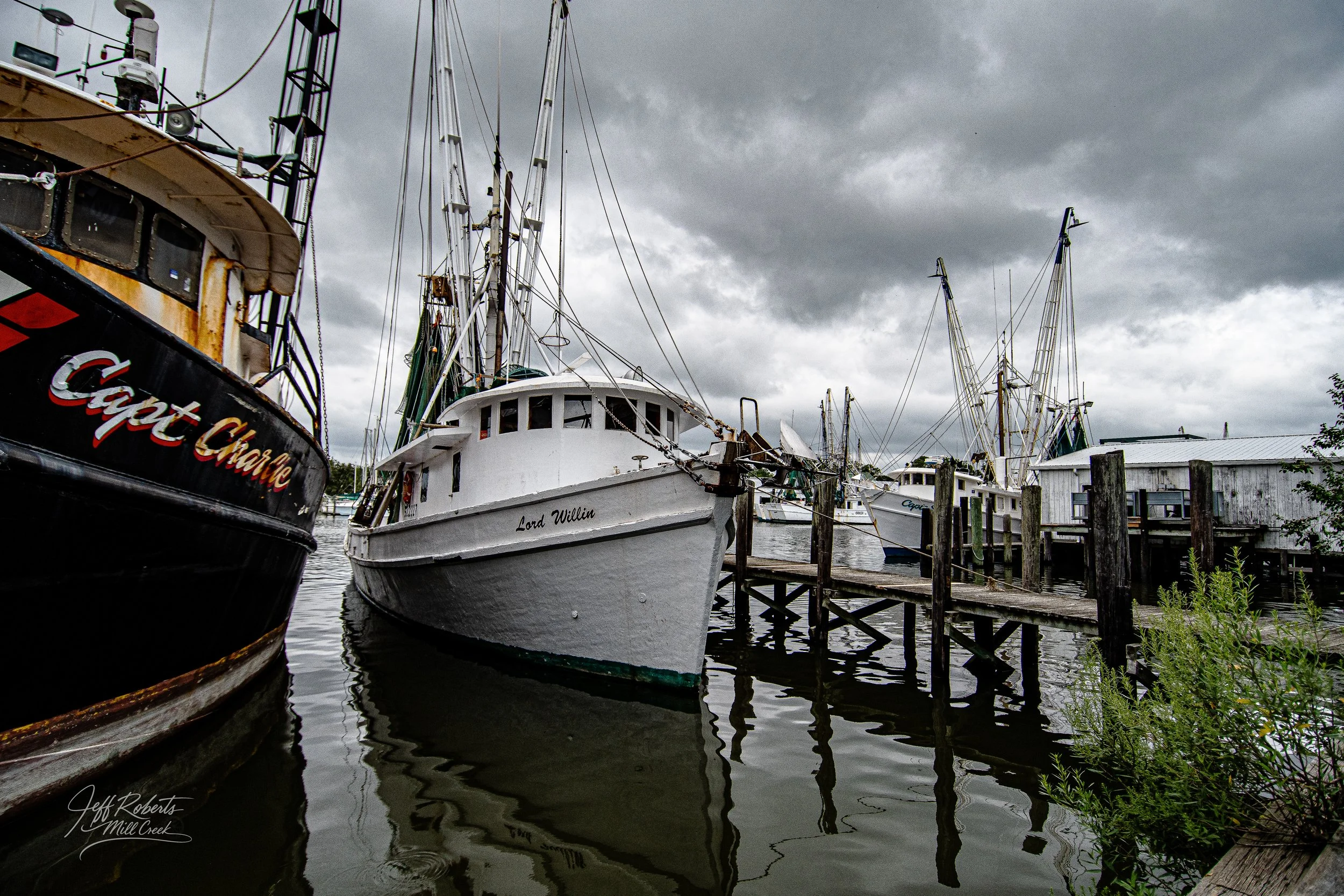 Several boats docked at a marina on a cloudy day, with one boat named Lord Willin in the foreground and a weathered wooden dock.