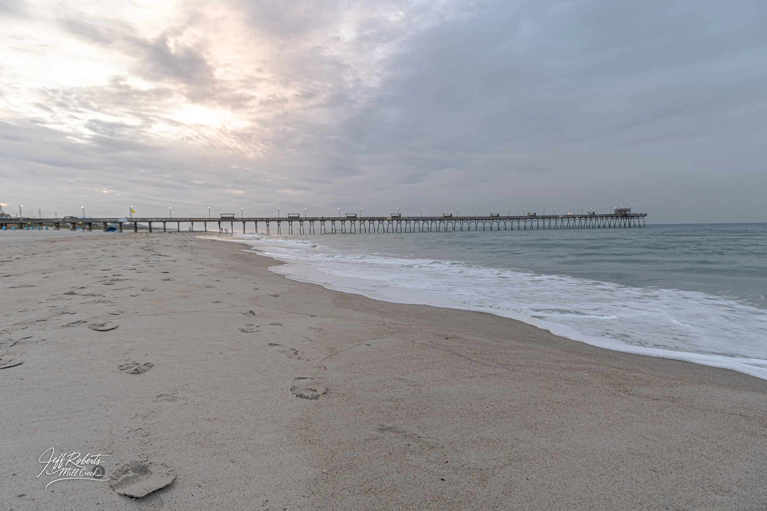Empty beach with footprints in the sand, calm ocean water, and a pier extending into the water under a cloudy sky.