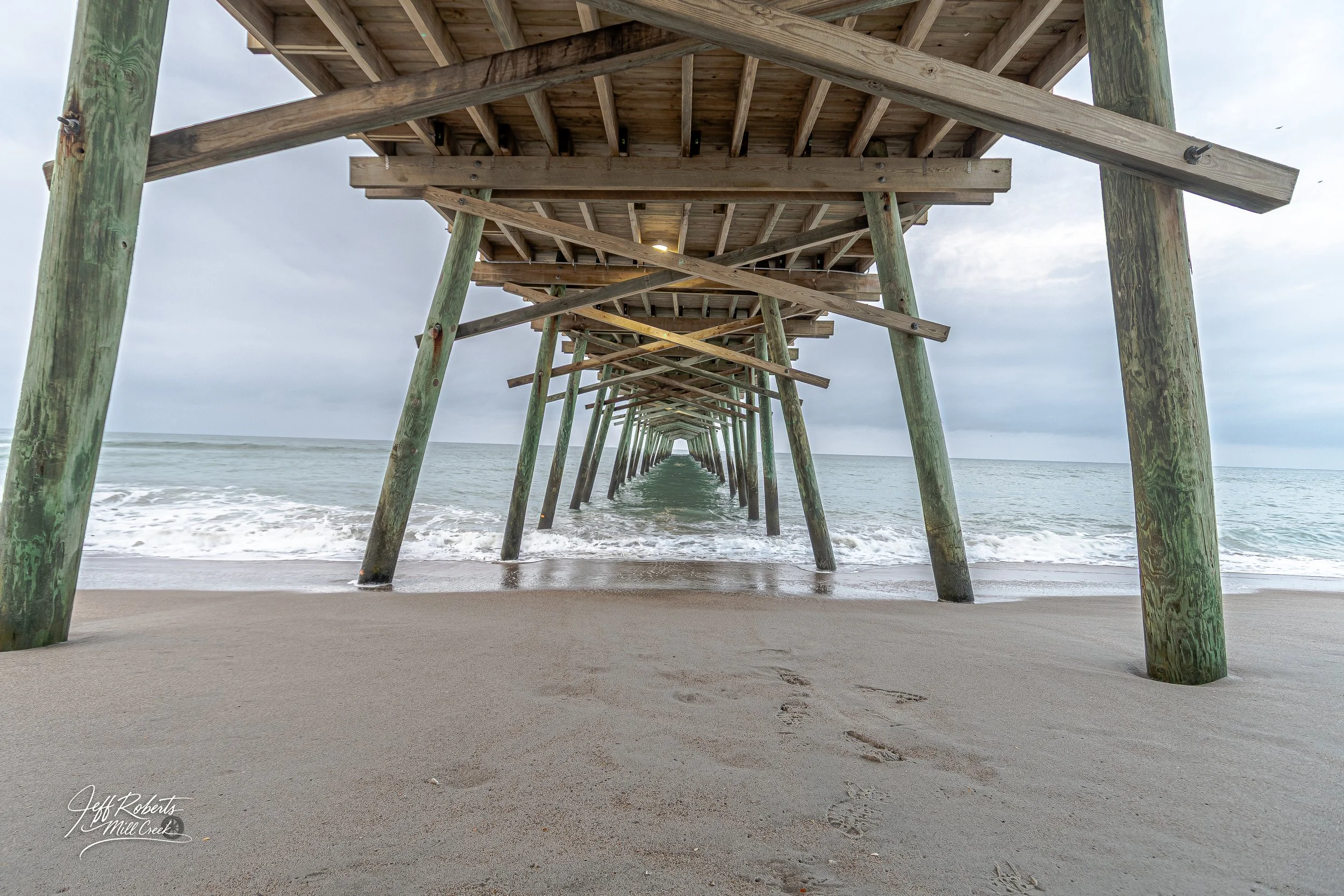 Underneath a wooden pier on a sandy beach with footprints, ocean waves, and cloudy sky