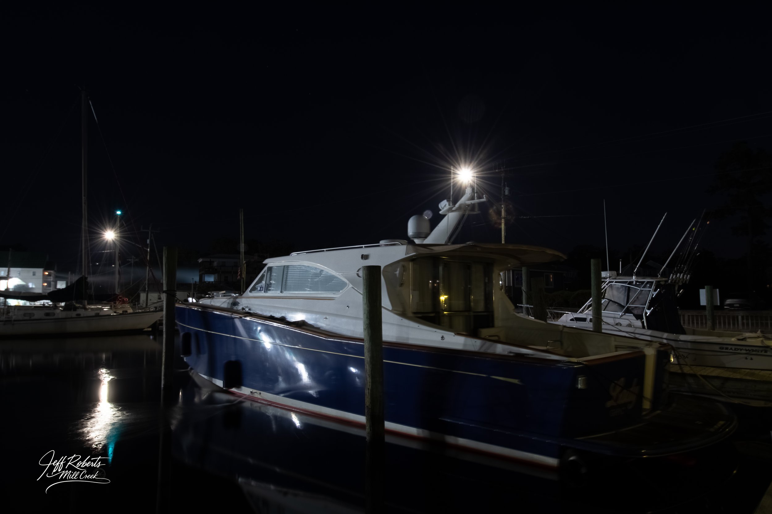 Nighttime scene at a marina with a boat docked, illuminated by a bright light on its mast, surrounded by other boats and docks, with dark water and a faint reflection of the boat.