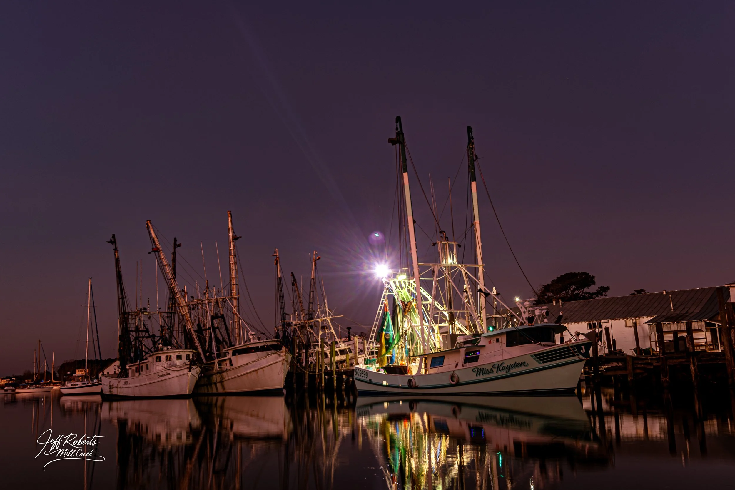Nighttime scene at a harbor with several anchored fishing boats, some with tall masts and rigging, reflected in calm water, illuminated by a bright light source, with a dark sky and some buildings along the dock.
