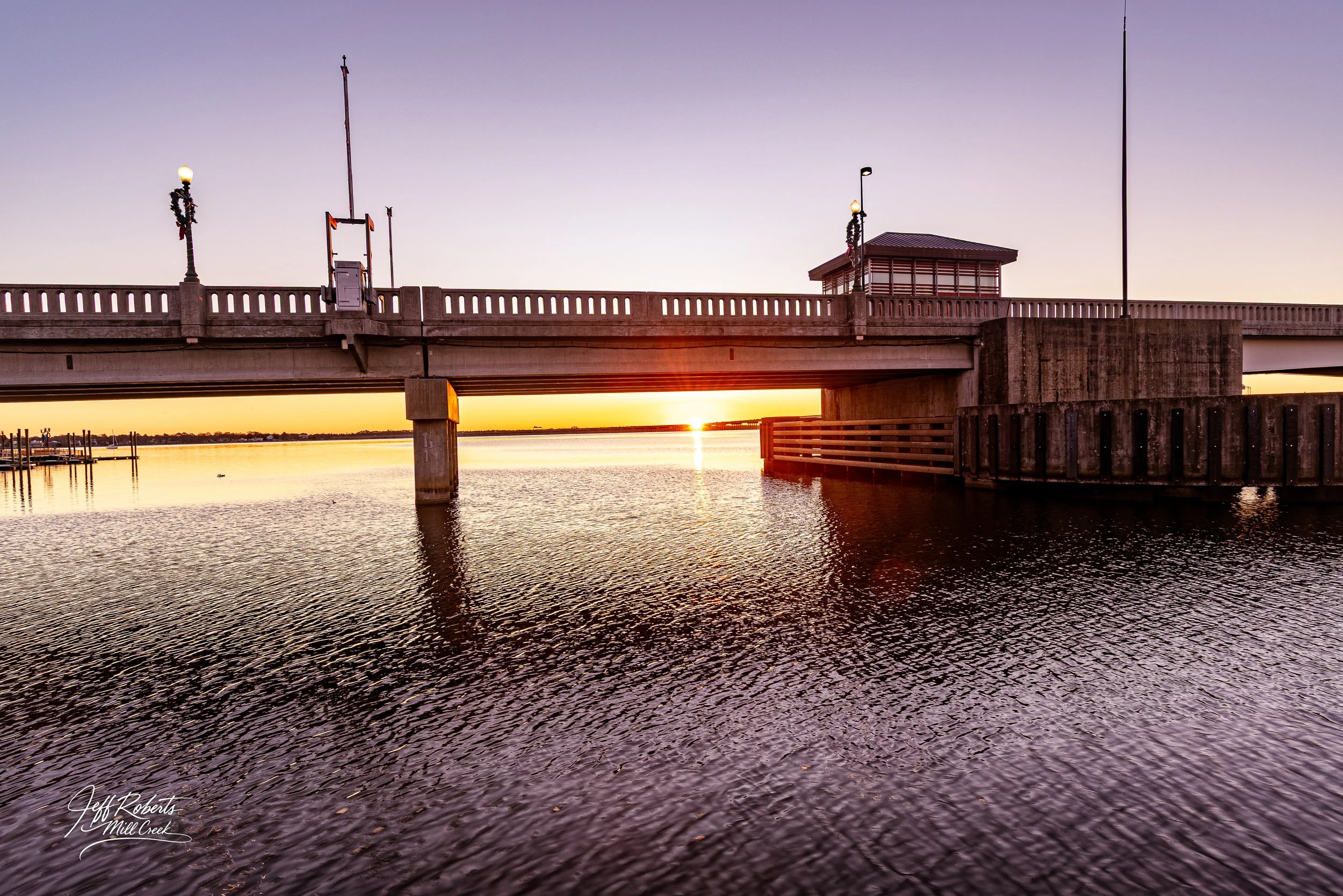 A bridge over a body of water during sunset, with the sun near the horizon casting an orange glow. The bridge has lamp posts decorated with wreaths and a small structure at one end.