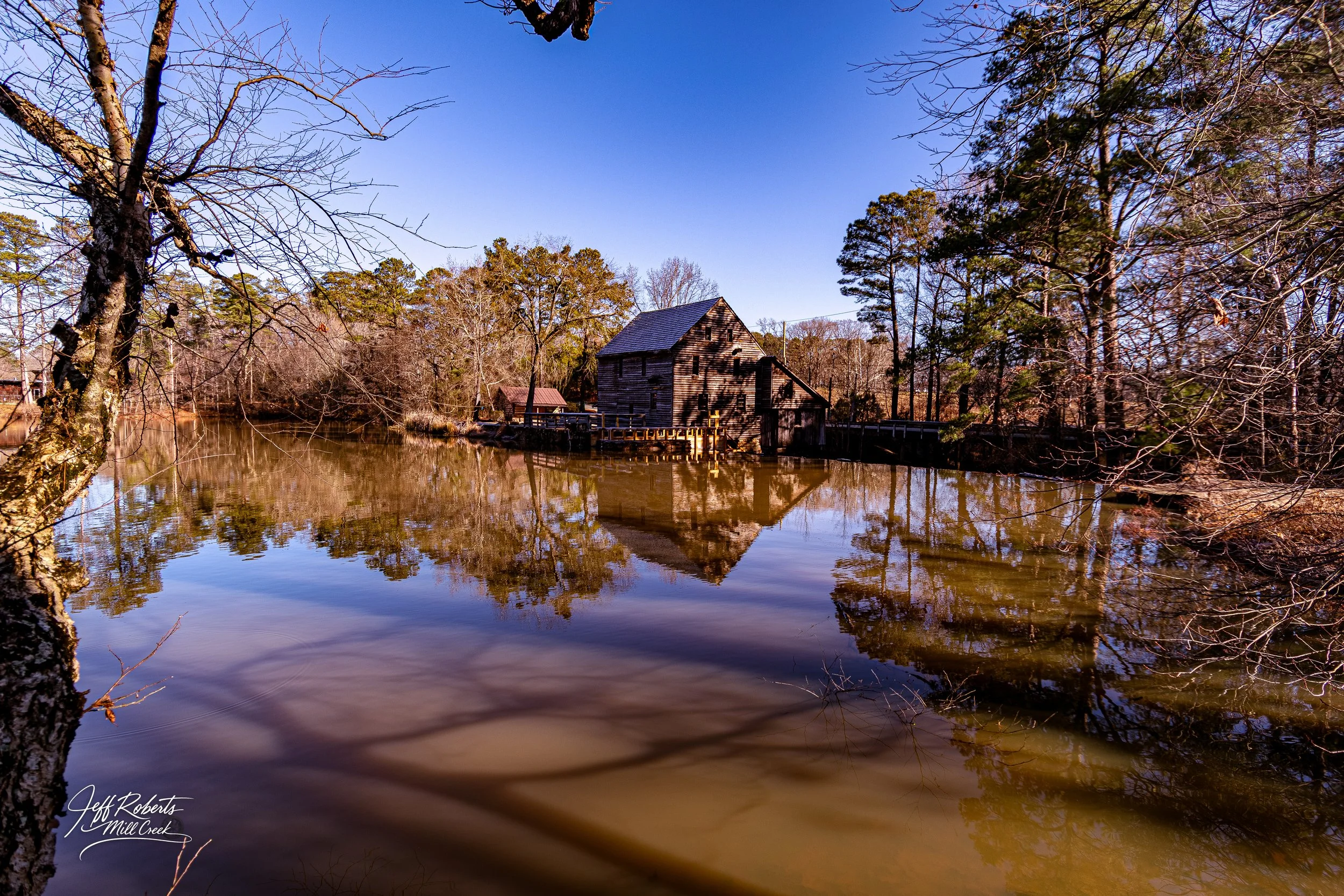 Old wooden mill house by a pond, surrounded by trees, with a clear blue sky overhead.
