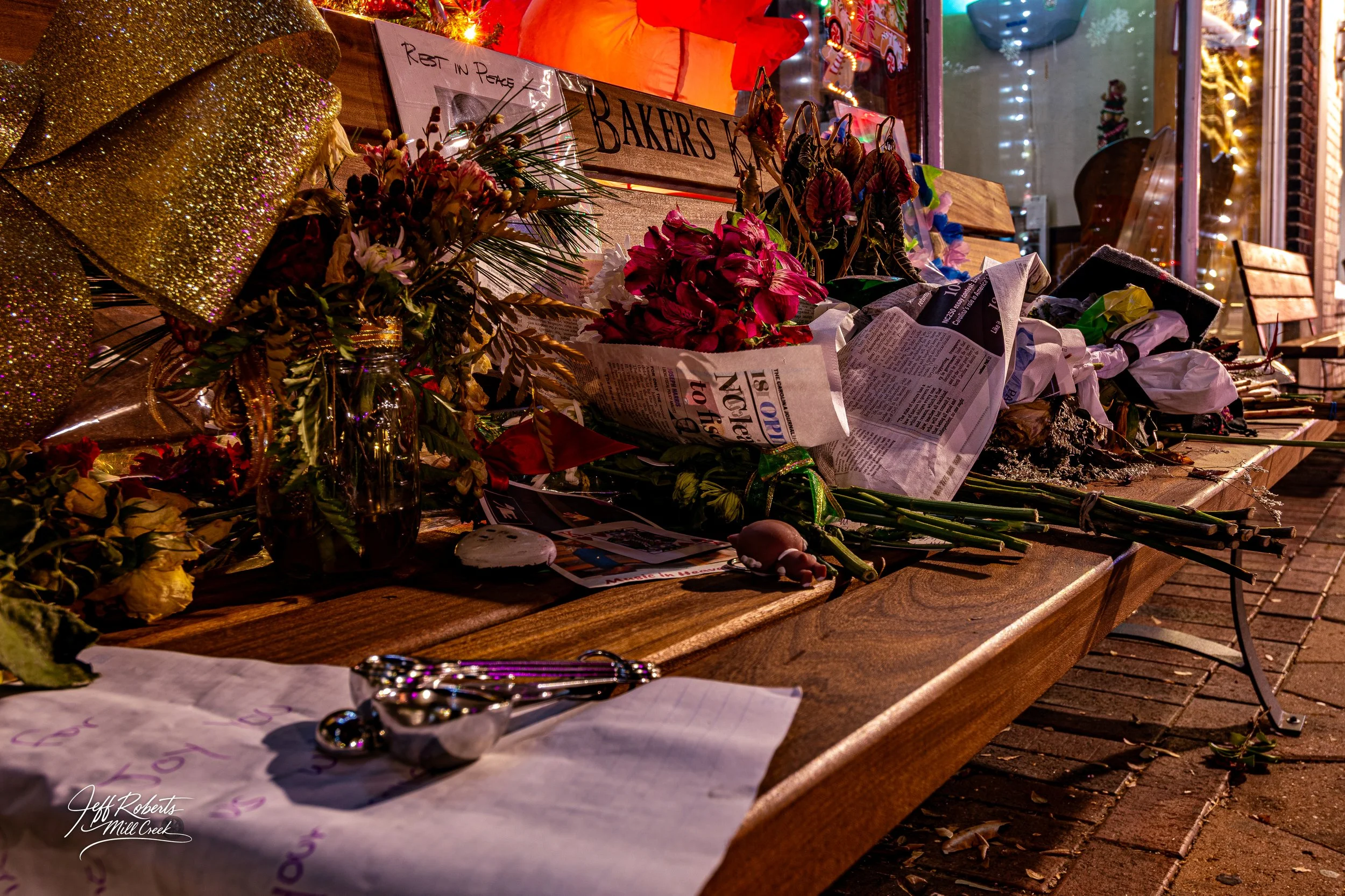 A memorial on a wooden bench for a recent death, containing flowers, newspapers, candles, and notes in a nighttime setting outside a storefront decorated with Christmas lights.