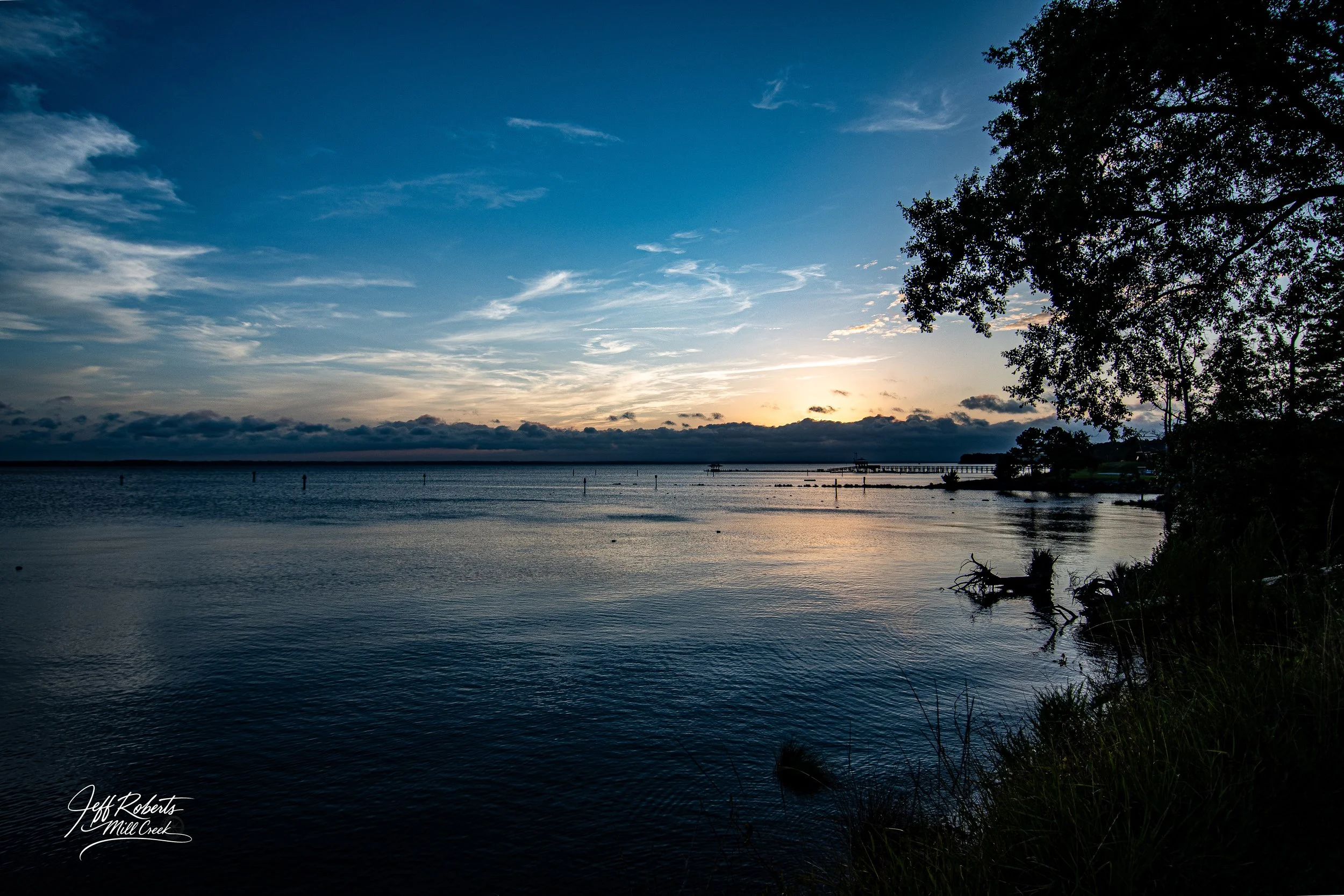 A serene body of water at sunset with blue skies and scattered clouds, silhouette of trees on the right, and a pier or dock in the distance.