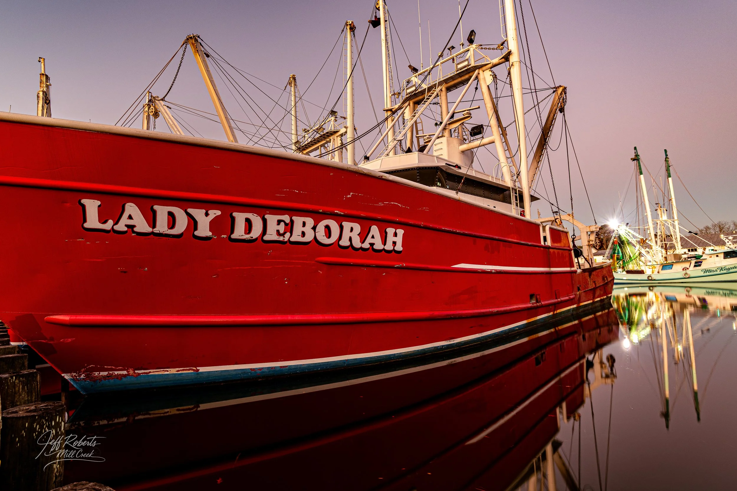 A red boat named Lady Deborah docked at a marina with its reflection visible in the water, several other boats are seen in the background during sunset or dusk.