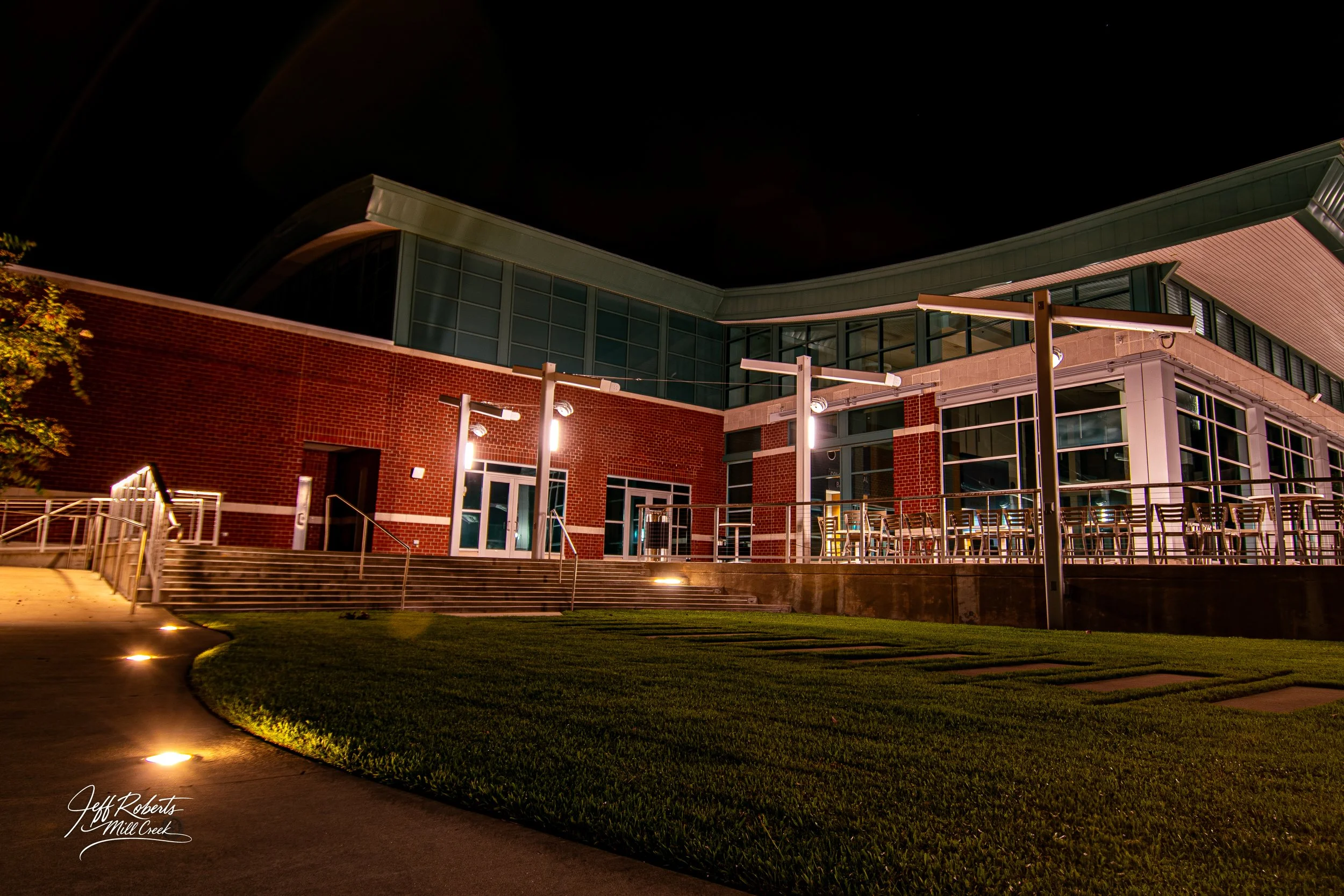 Night view of a modern building with brick and glass exterior, outdoor tables and chairs on a patio, and pathway lighting.