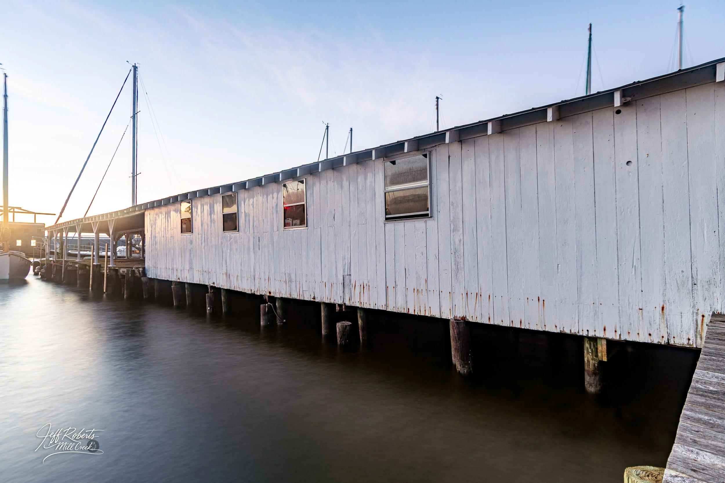 White weathered building on stilts along a waterfront with boats docked nearby, early morning or late afternoon sunlight