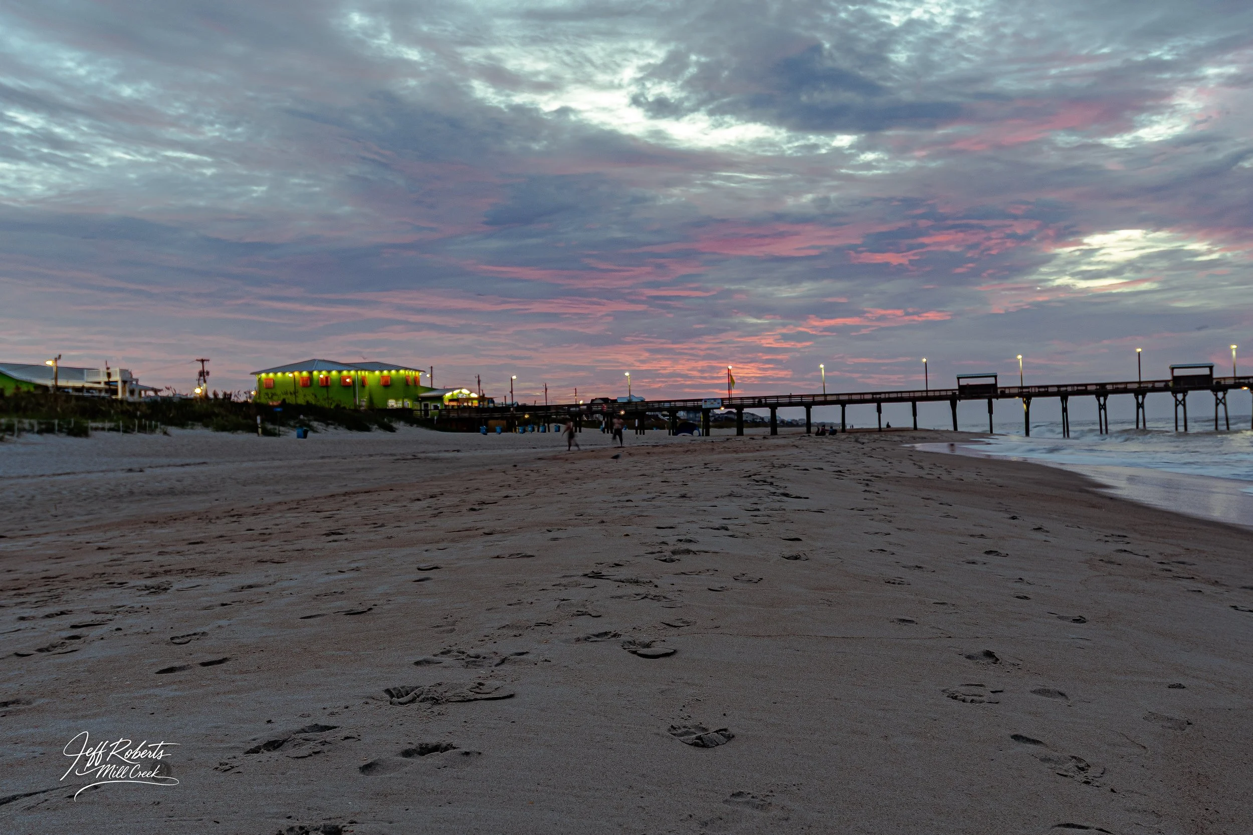 A beach at sunset with footprints on the sand, a pier extending into the ocean, and a brightly lit green building near the pier, under a partly cloudy sky with pink and blue hues.