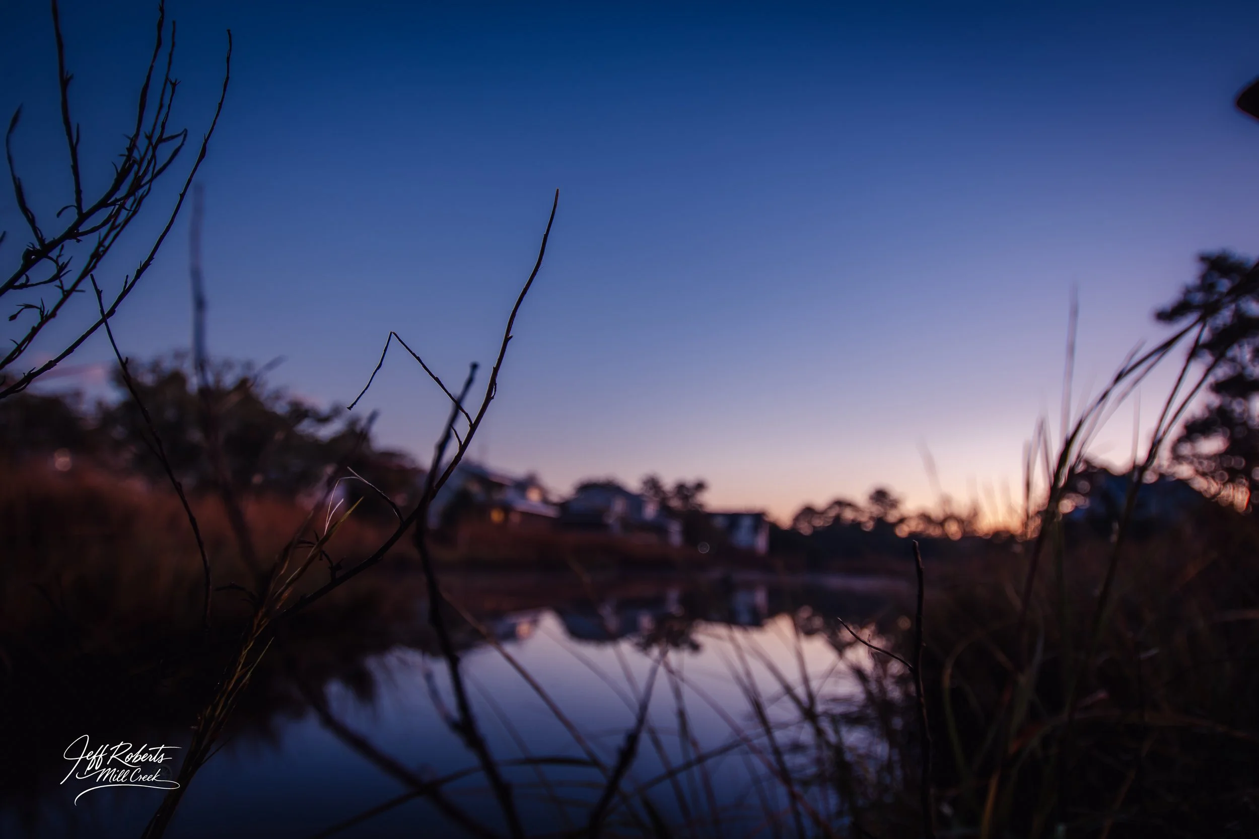 A tranquil scene at dusk or dawn showing a river with reflections, silhouetted branches in the foreground, and houses or buildings in the background under a gradient sky transitioning from purple to light blue.